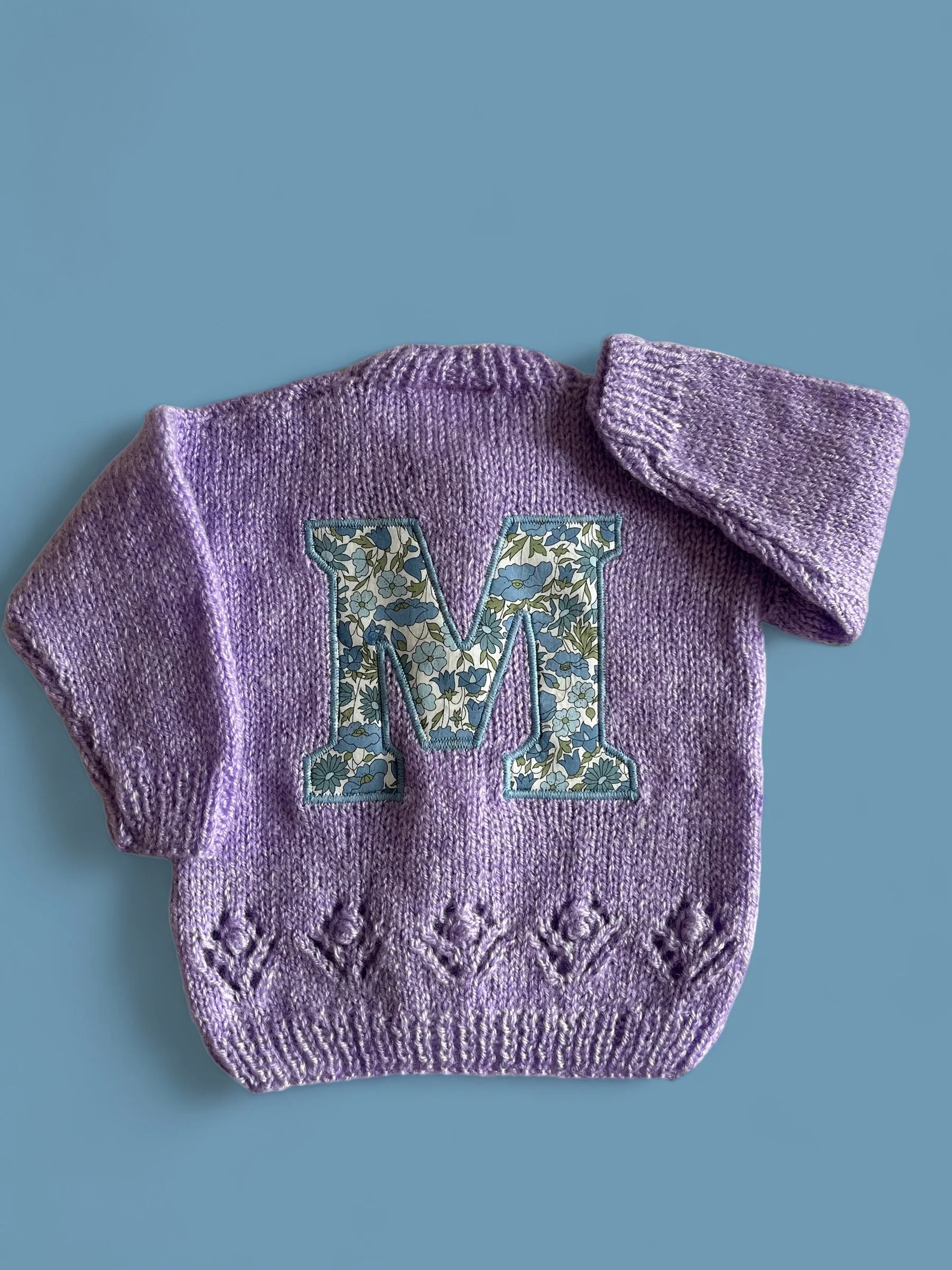 A lavender knitted sweater with a large, floral patterned letter 'M' on the front and decorative knitting at the bottom, lying on a light blue surface.