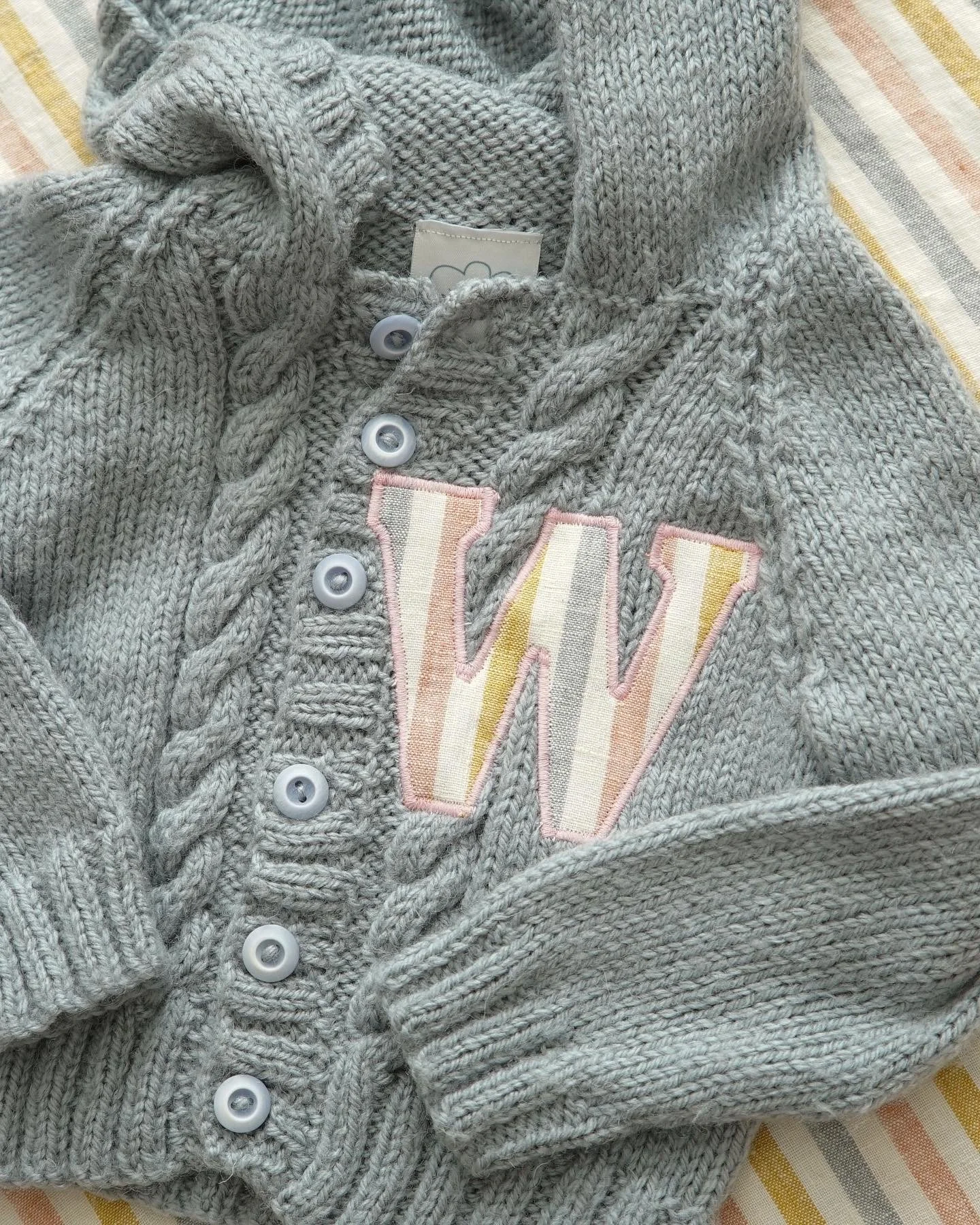Gray knitted sweater with a hood and "W" patch, featuring cable knit pattern and buttons, on striped background.