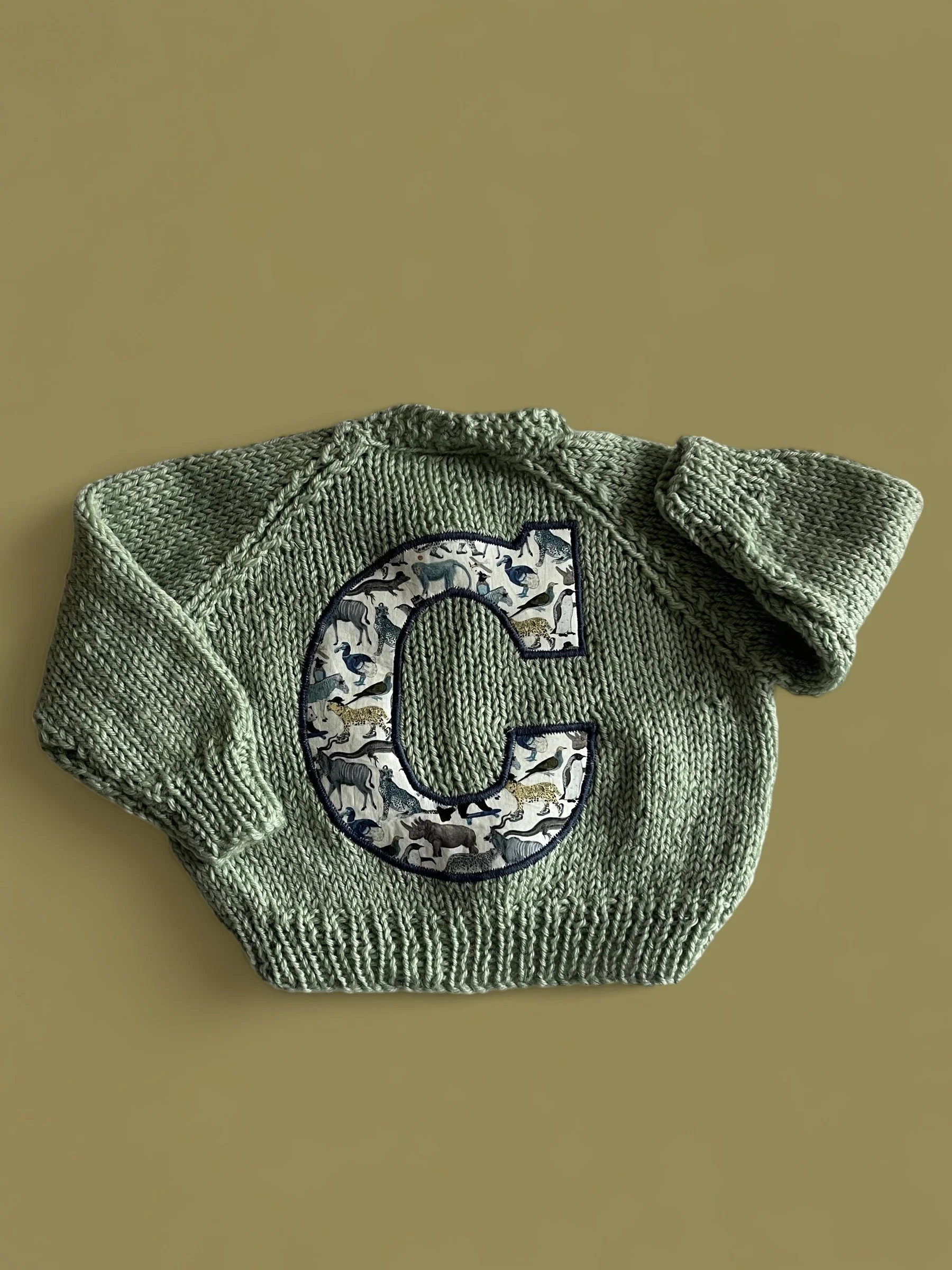 Green knitted baby sweater with a large letter 'C' patch featuring animal illustrations.