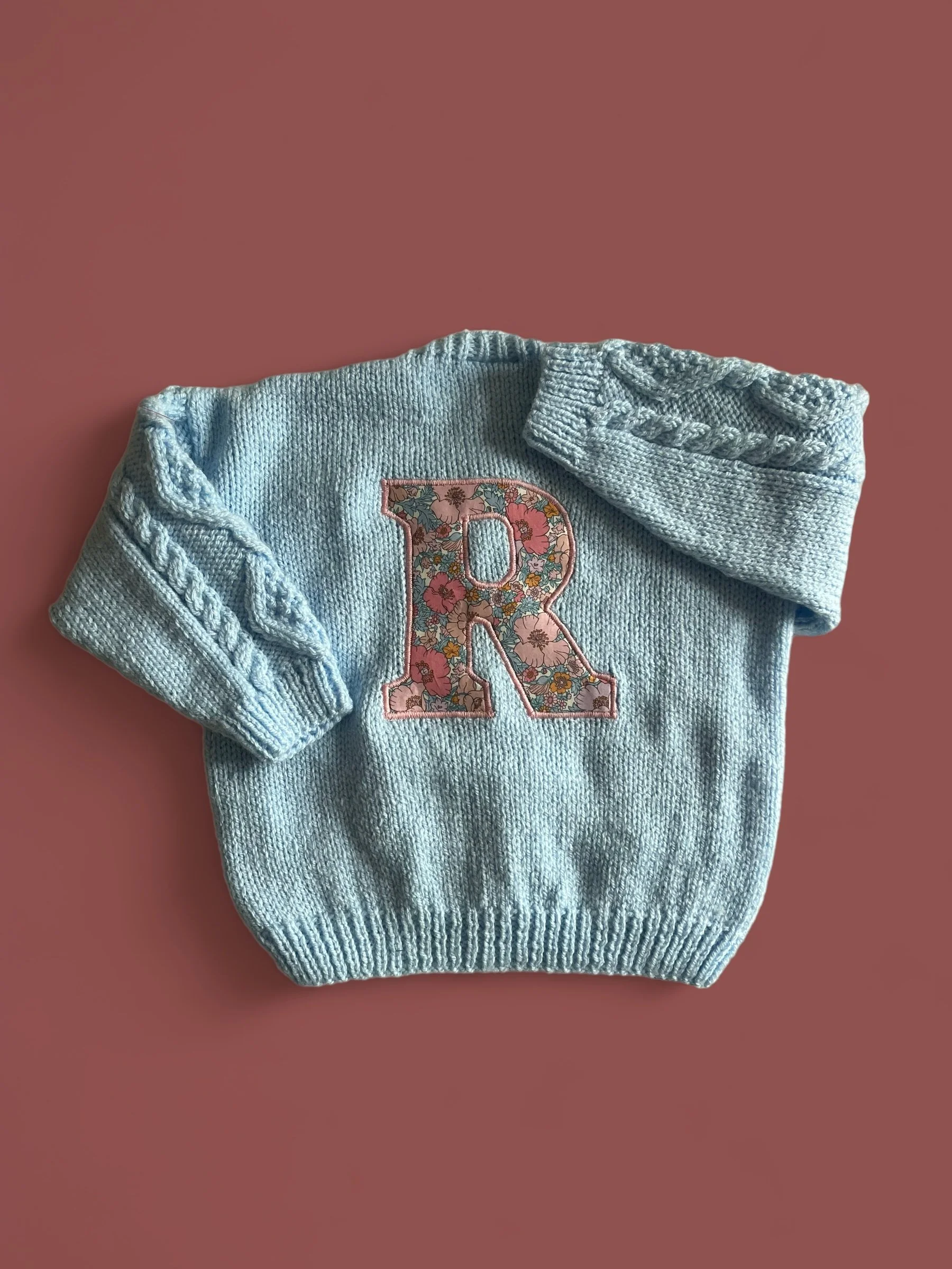 Baby blue knitted sweater with a floral embroidered letter R on the front, laid on a pink background.