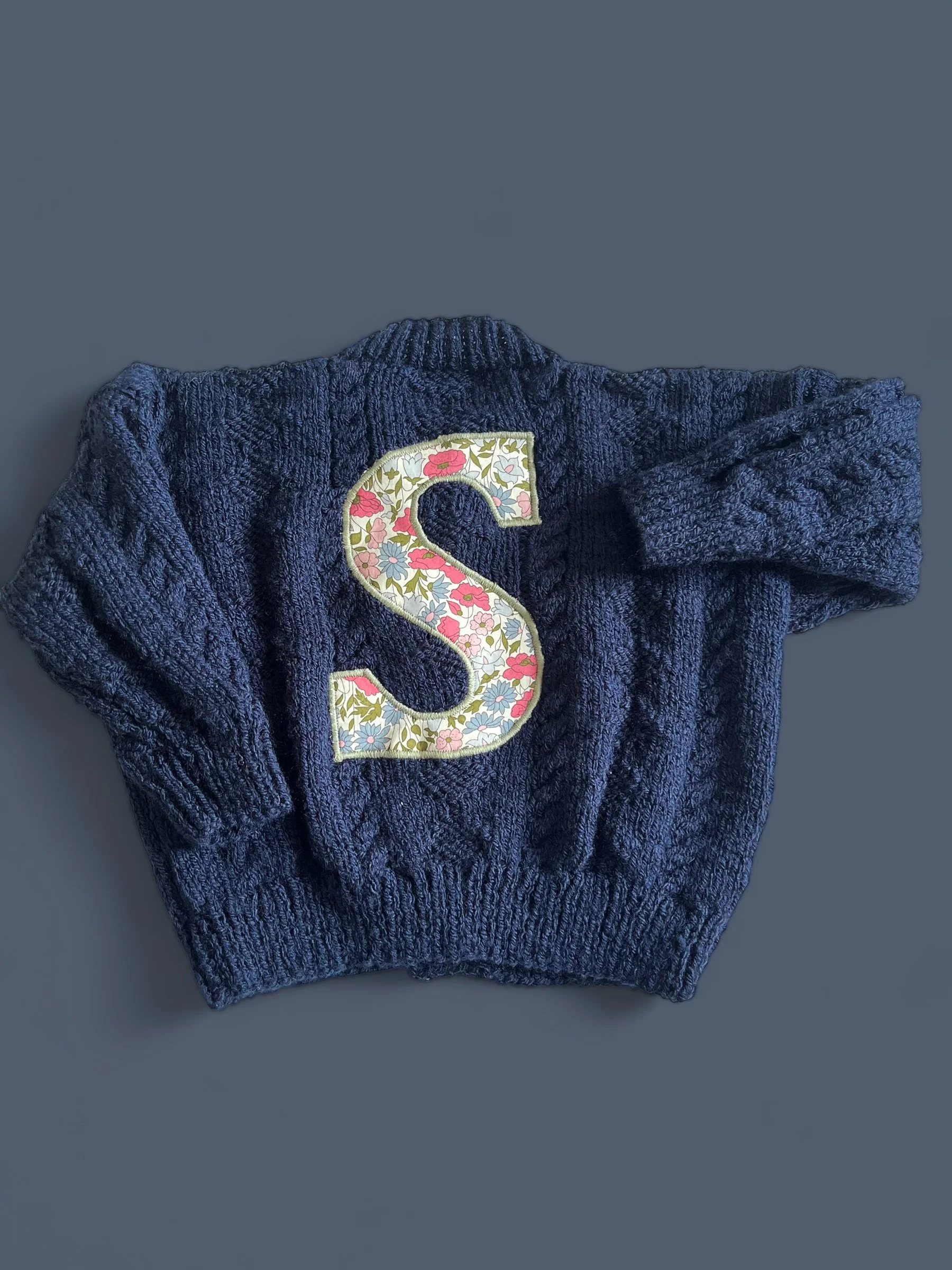 A navy blue knitted sweater with a large letter 'S' patch featuring a floral pattern, laid against a gray background.