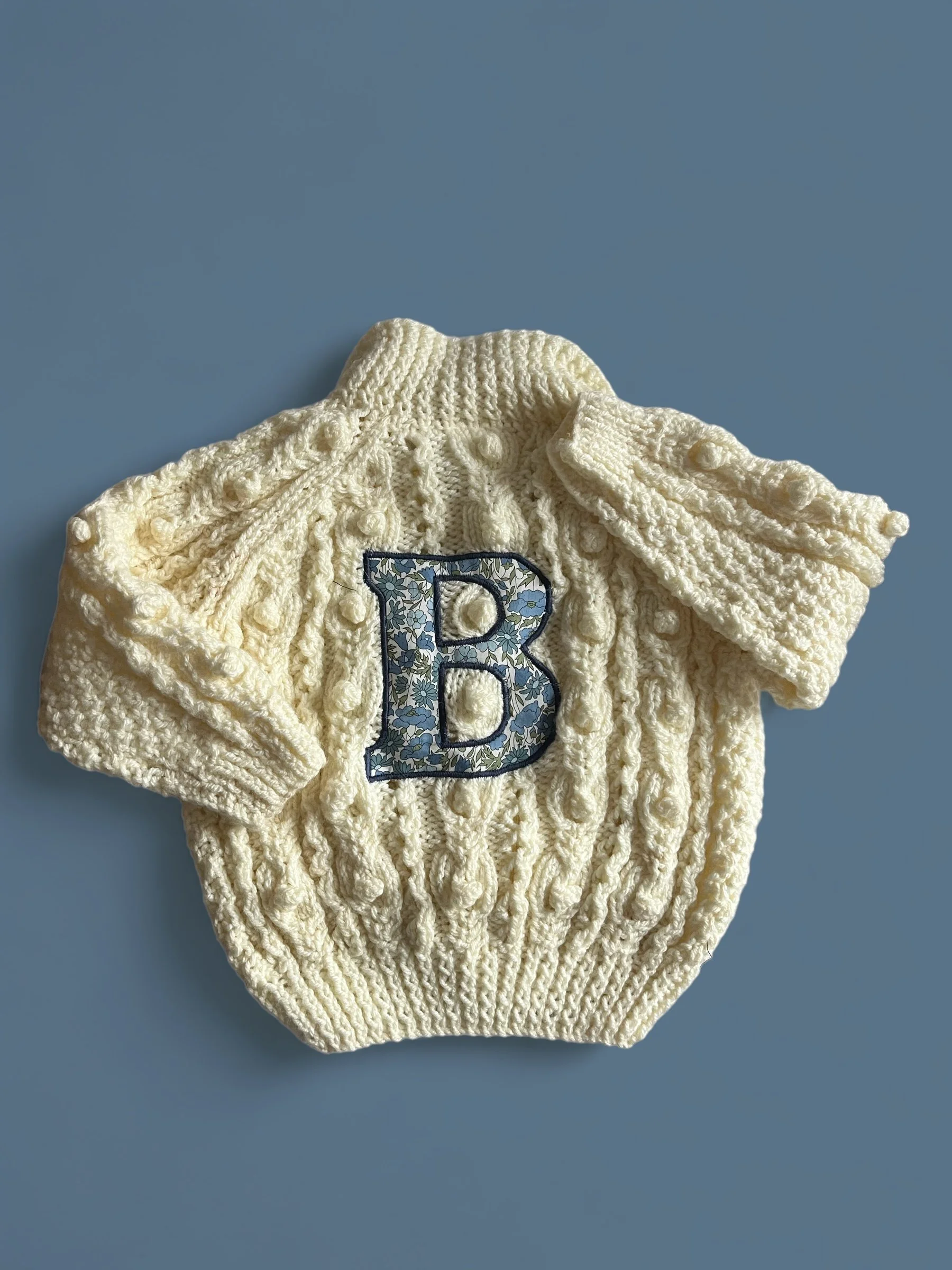 Cream-colored knitted baby sweater with a blue floral letter B patch on the front, laid flat on a light blue background.