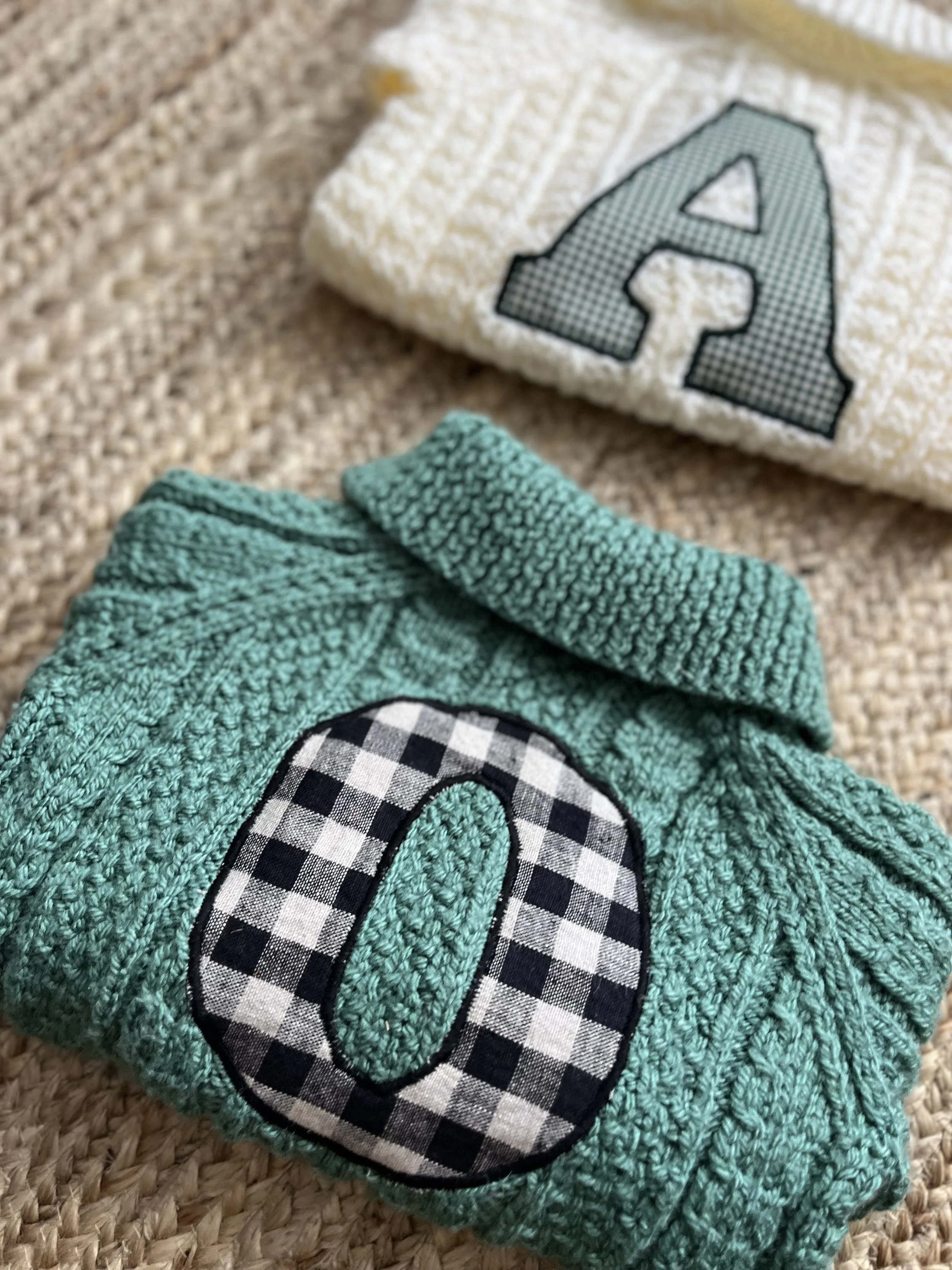 Two folded cable knit sweaters with letter patches, one white with an 'A' and one green with an 'O' on a textured surface.