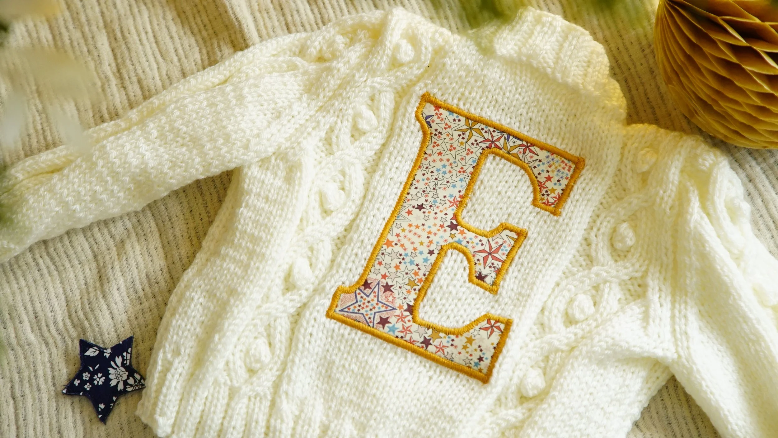 A white knitted sweater with a large embroidered letter 'E' in colorful star pattern and a yellow border, resting on a textured surface with a small star-shaped patch nearby.