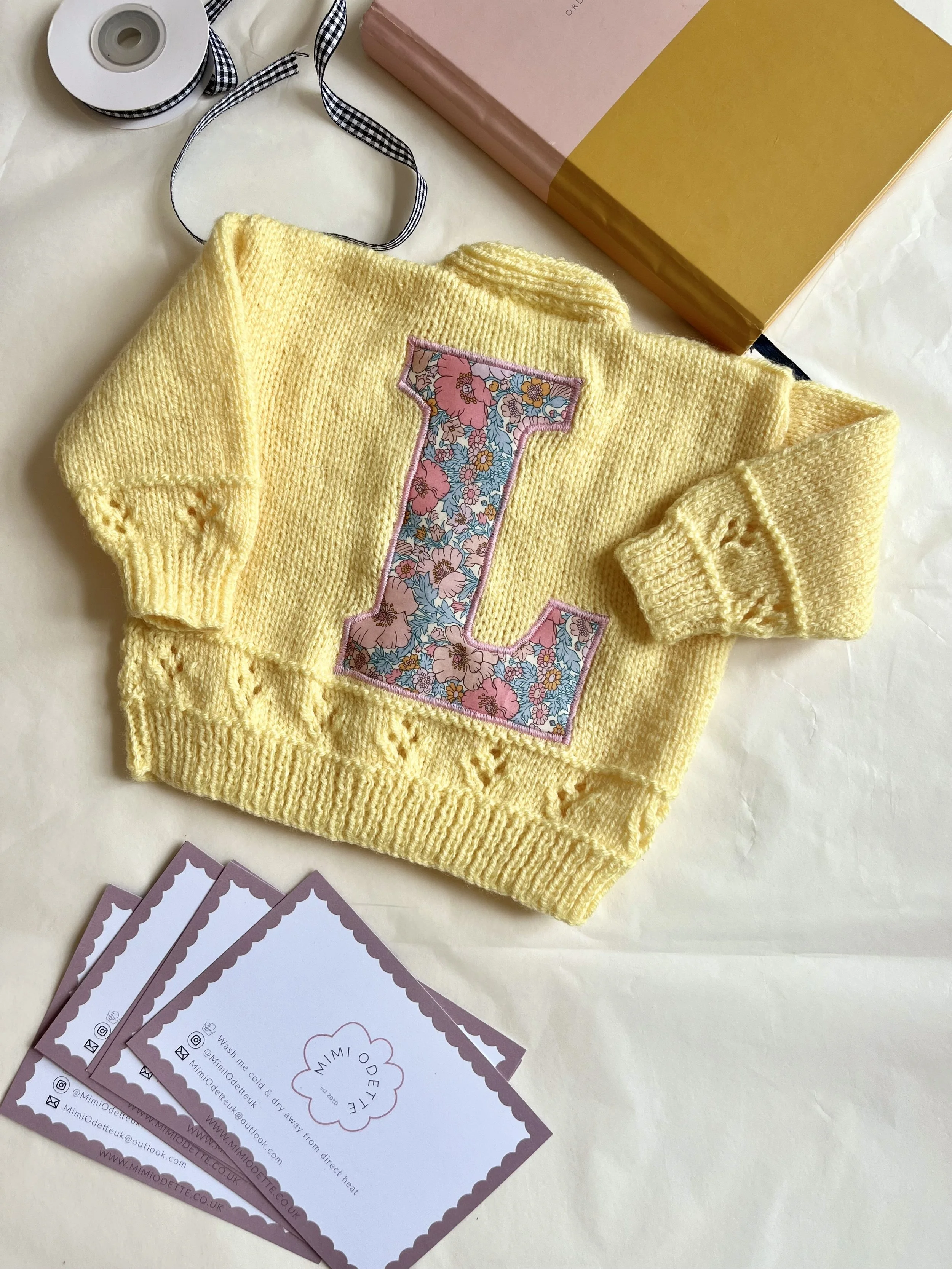 Yellow knitted sweater with a floral letter 'L' patch on the front, next to business cards, a pink and brown box, a roll of ribbon, and a piece of white paper.