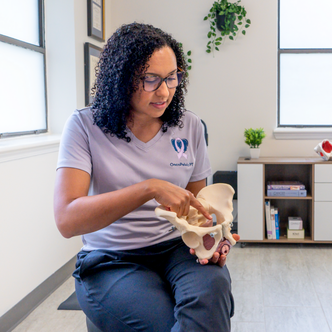 Dr Alex Hill, board certified physical therapist in pelvic & women's health and oncology physical therapy, is sitting in her clinic treatment room holding a pelvis teaching a patient