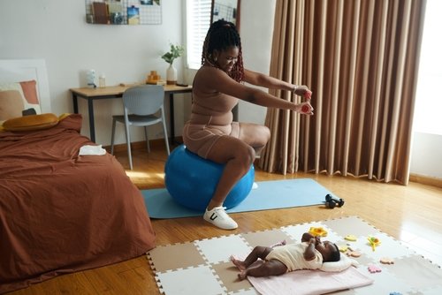 Black mother sitting on blue exercise ball exercising, smiling at her baby playing on the floor