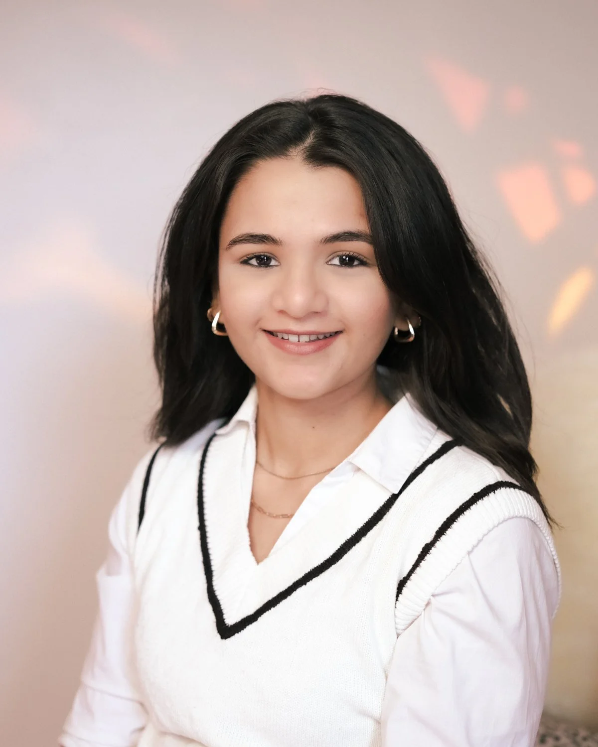 A young woman with dark hair, wearing a white shirt and a white vest with black stripes, smiling at the camera against a neutral background.