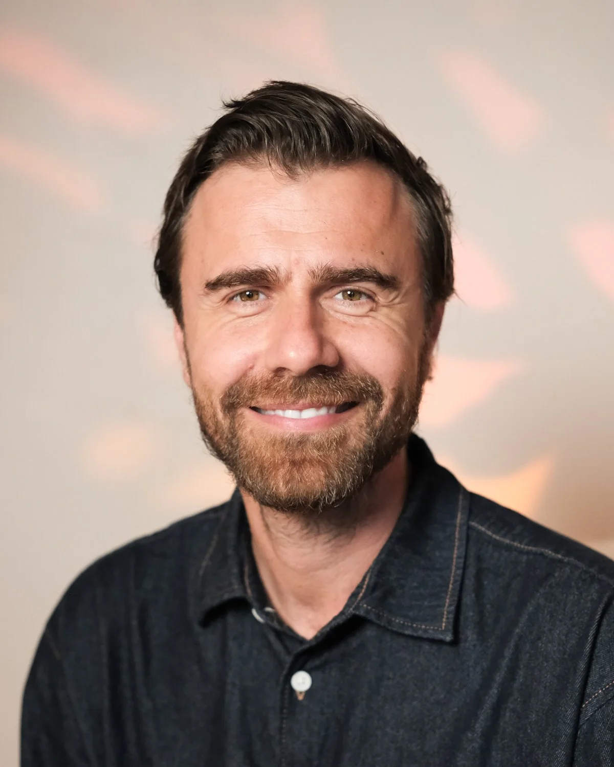 A smiling man with brown hair and a beard, wearing a dark shirt, standing against a neutral background.