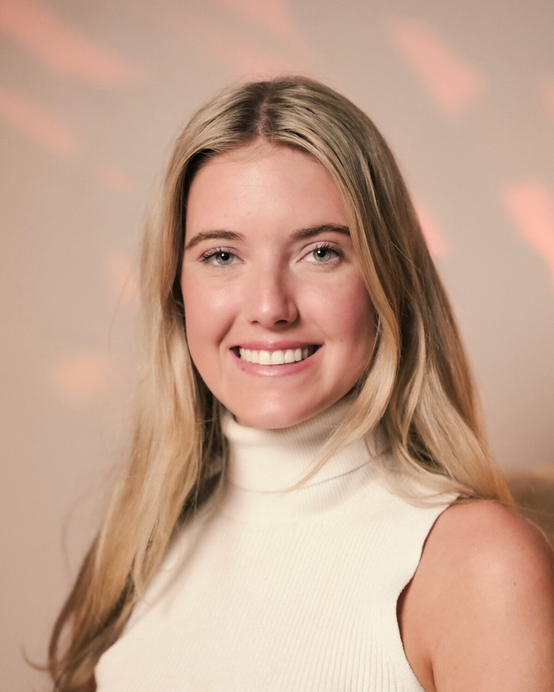 A young woman with blonde hair, blue eyes, and a white sleeveless turtleneck top smiling at the camera, with a blurred background featuring pink and yellow abstract light patterns.