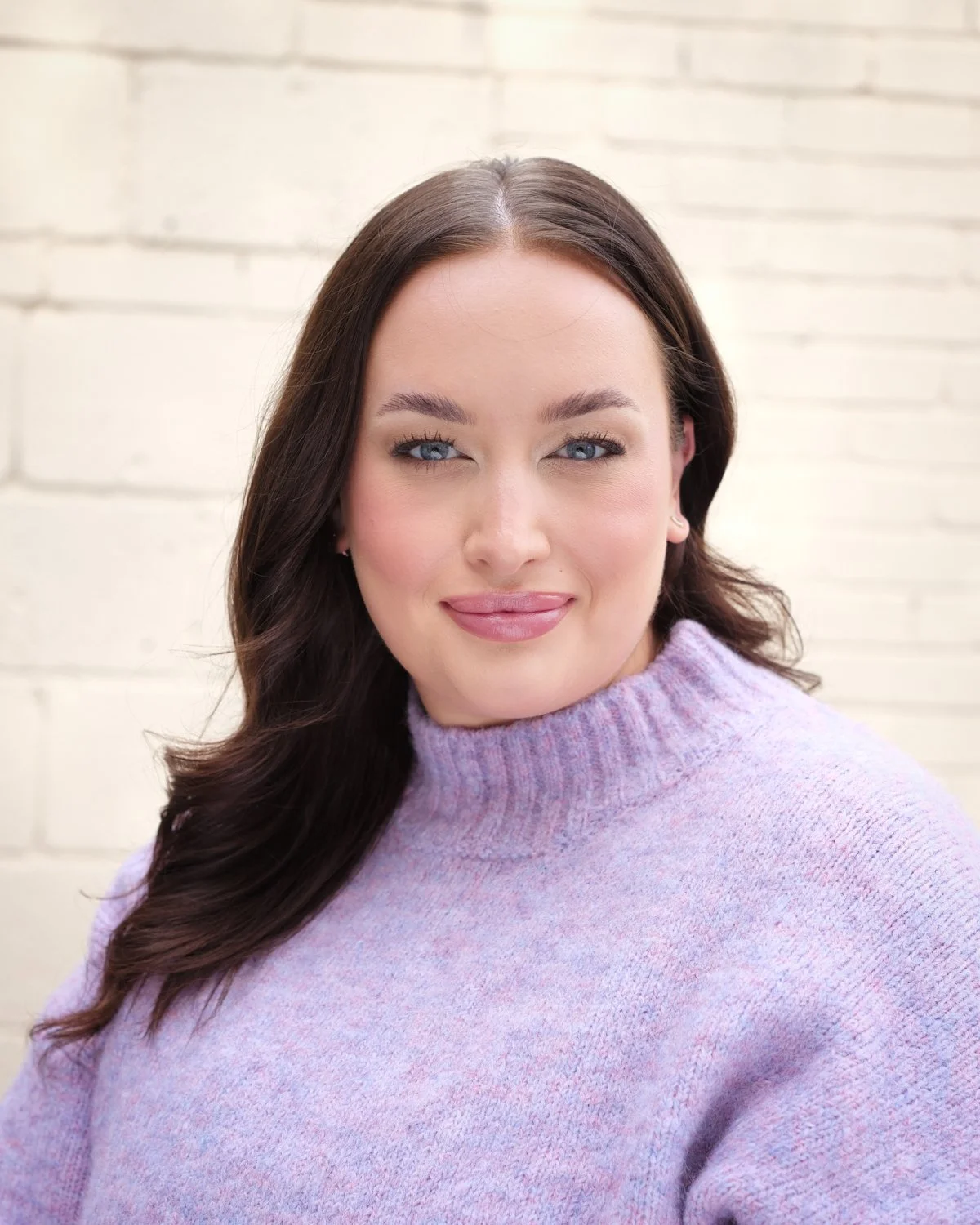 A woman with long brown hair, blue eyes, and light pink lipstick, wearing a lavender sweater, smiling in front of a white brick wall.