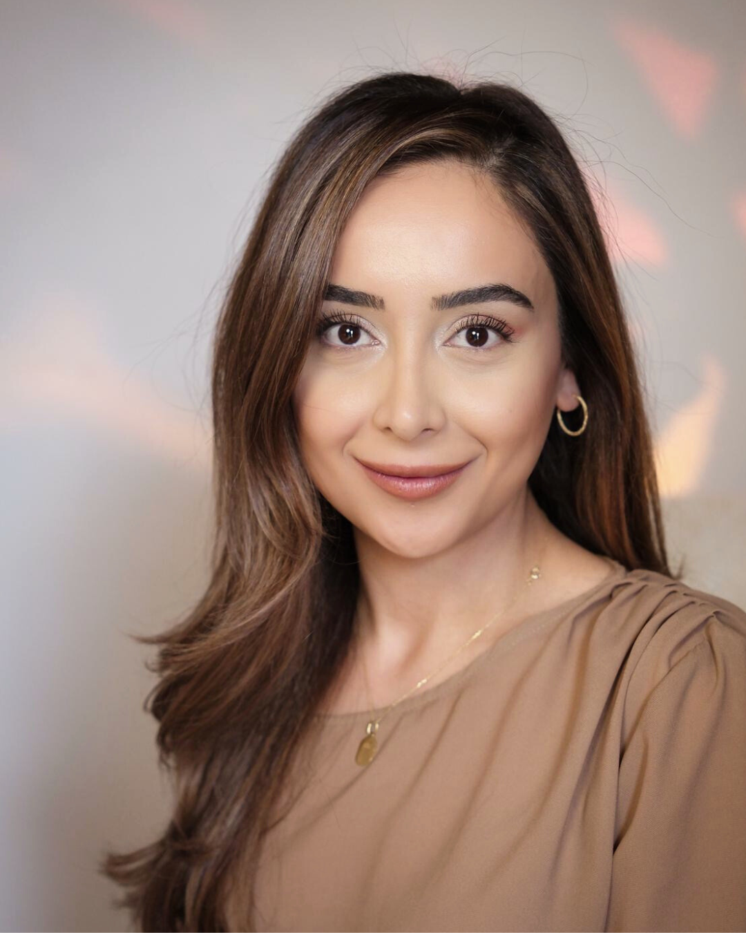 A woman with light skin, long wavy brown hair, and brown eyes, smiling, wearing gold hoop earrings and a beige top, against a blurred colorful background.