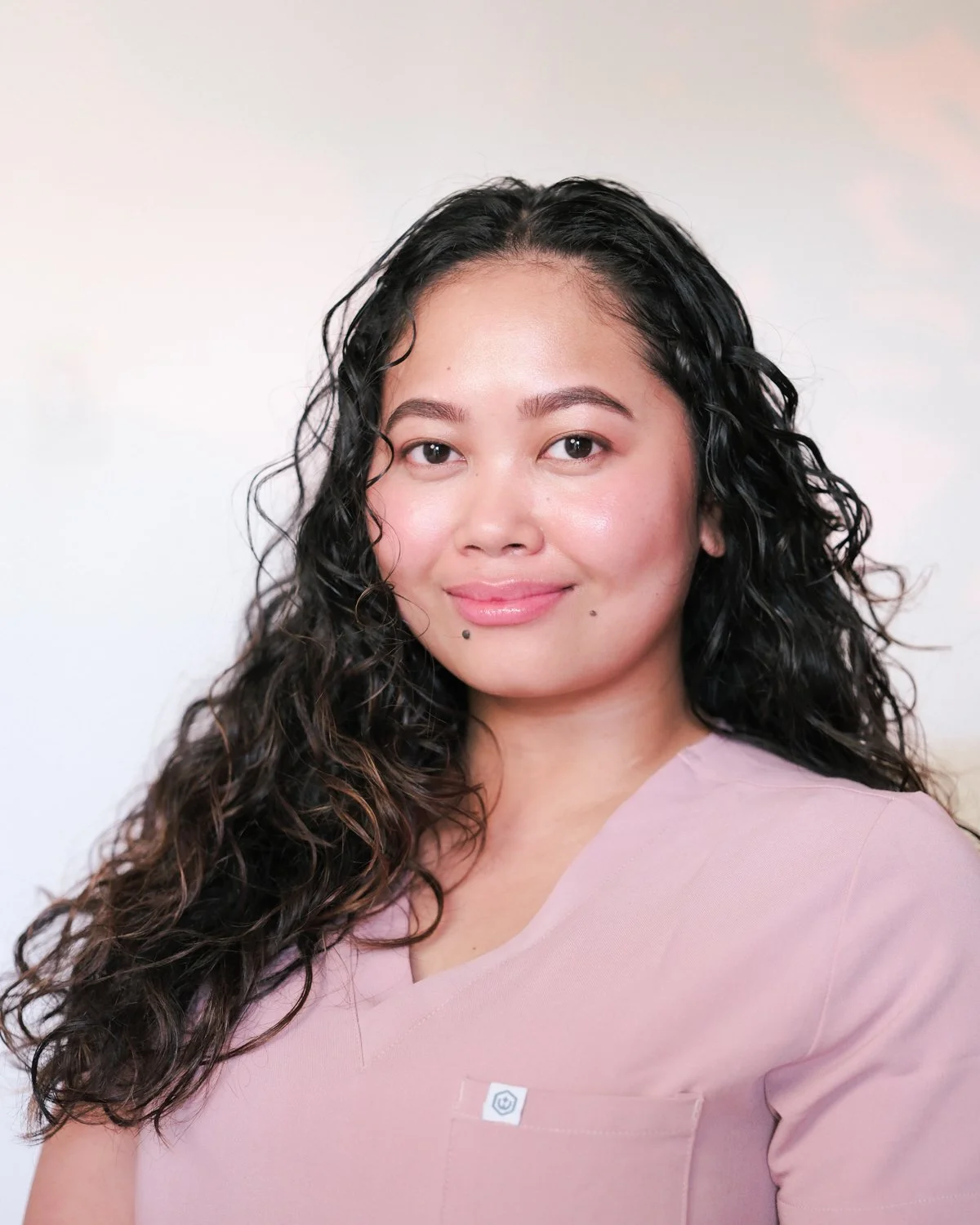 A woman with curly black hair and light skin wearing a light pink top, smiling softly against a plain light-colored background.