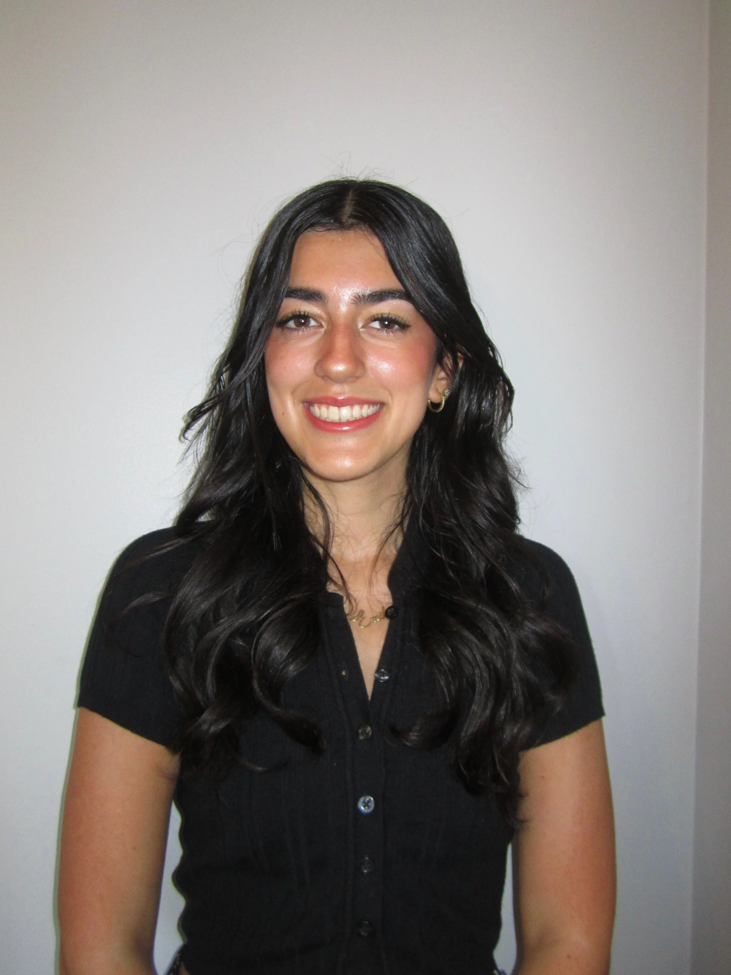 A young woman with long dark hair, smiling, wearing a black button-up shirt, standing against a plain white wall.