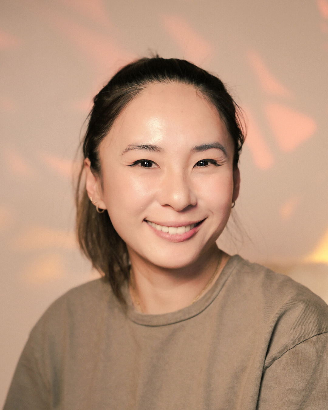 A woman with dark hair tied back, smiling, wearing a beige shirt, with a soft pink and yellow background.