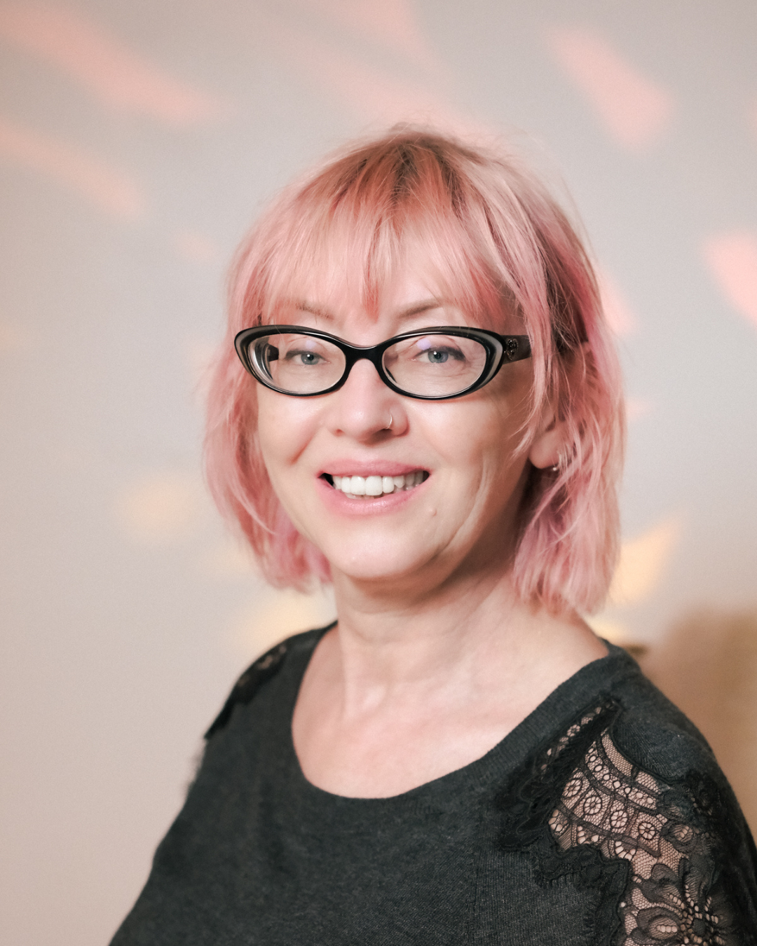 A woman with pink hair, black glasses, and a nose ring smiling at the camera, wearing a black top with lace details on the shoulders.