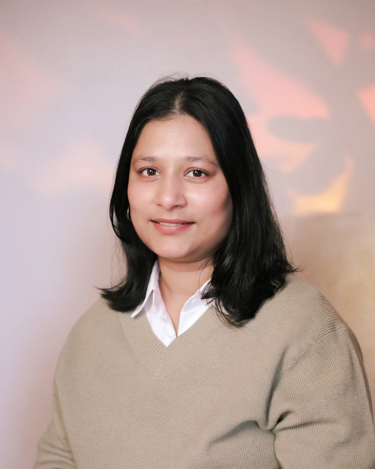 A woman with shoulder-length black hair, wearing a white collared shirt and beige sweater, smiling softly at the camera against a neutral background.