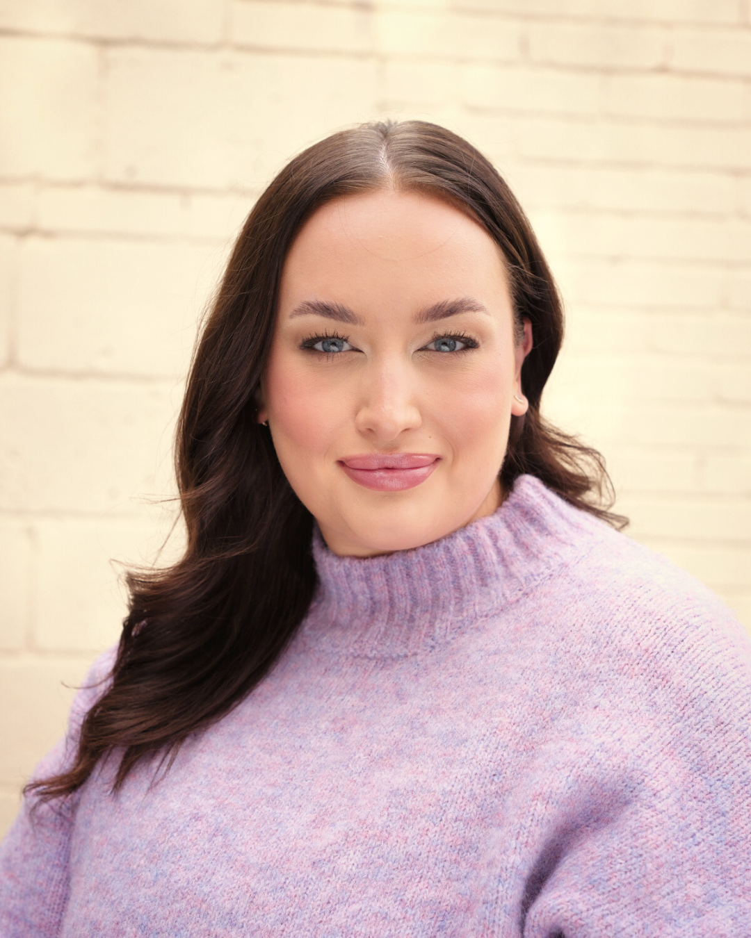 A woman with long brown hair, blue eyes, and light pink lipstick, wearing a lavender sweater, smiling in front of a white brick wall.