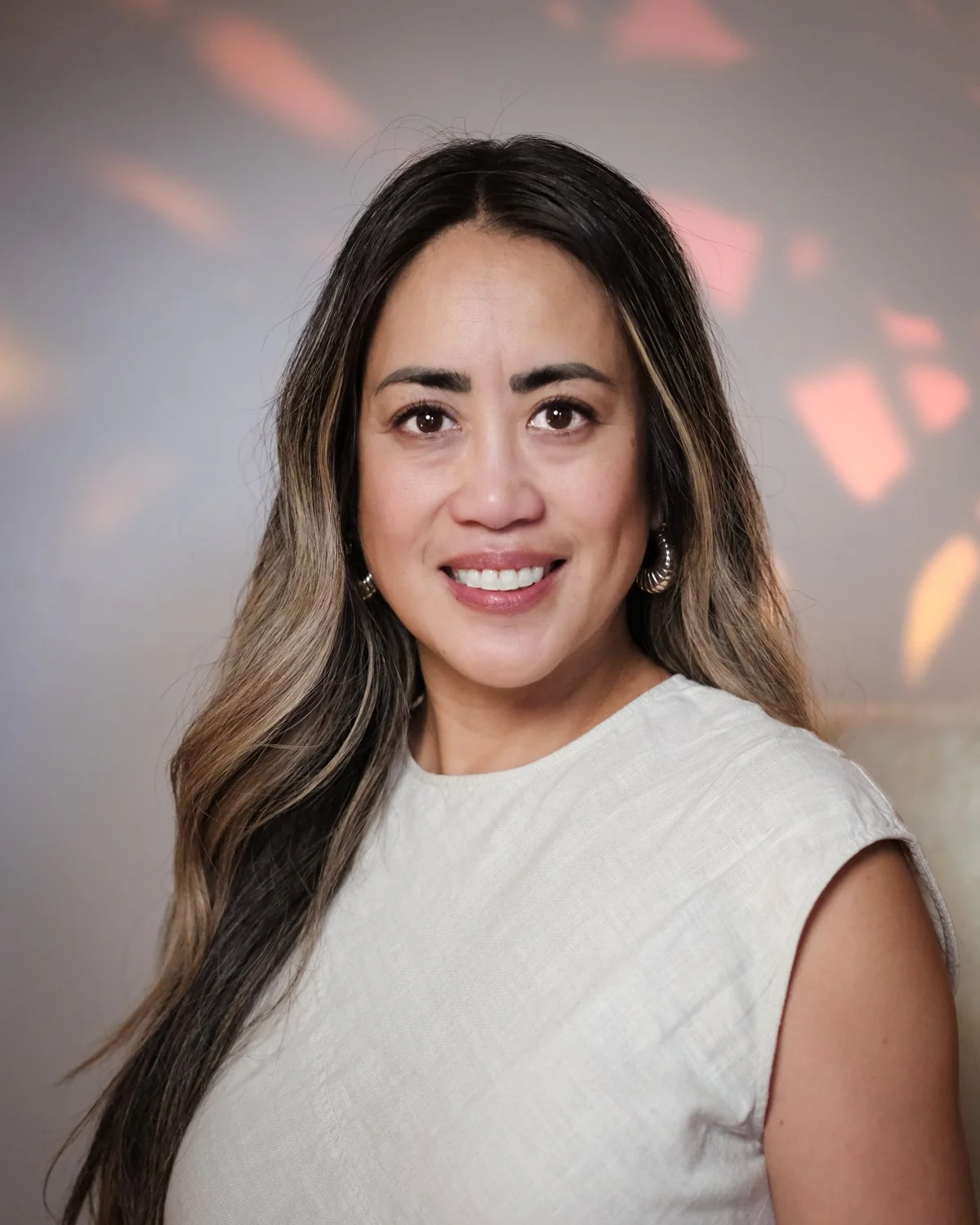 A woman with long wavy hair and a light-colored blazer stands in front of a white background with a large green monstera leaf to her side.