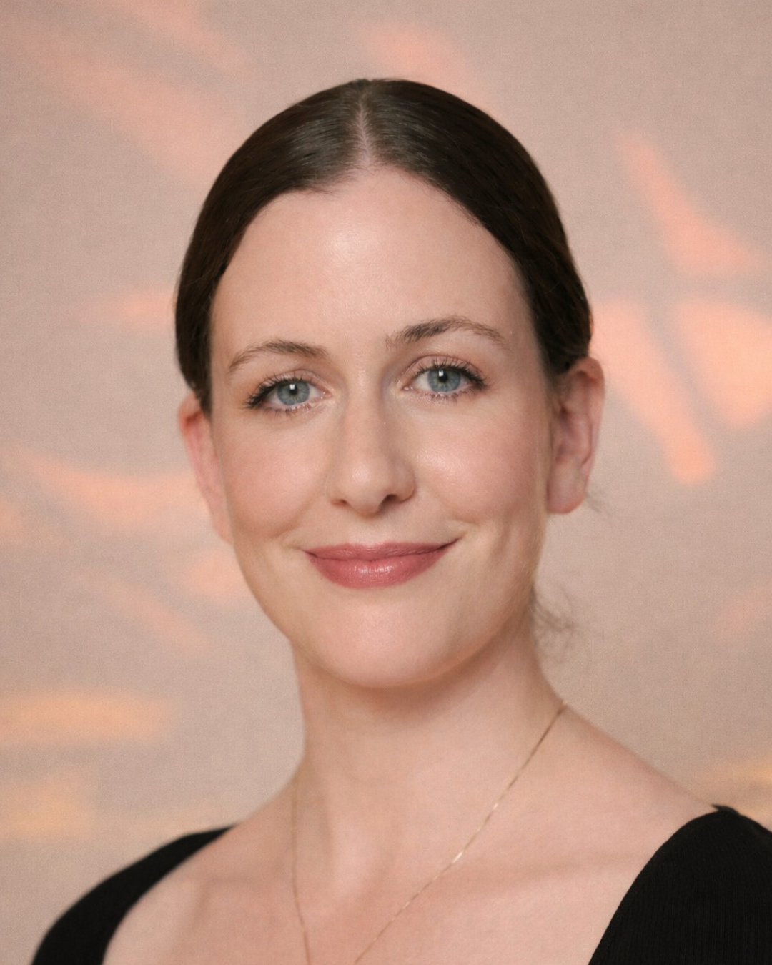 A woman with dark hair, dressed in a black sleeveless top, standing indoors with large green leaves of a Monstera plant beside her, against a light-colored wall.