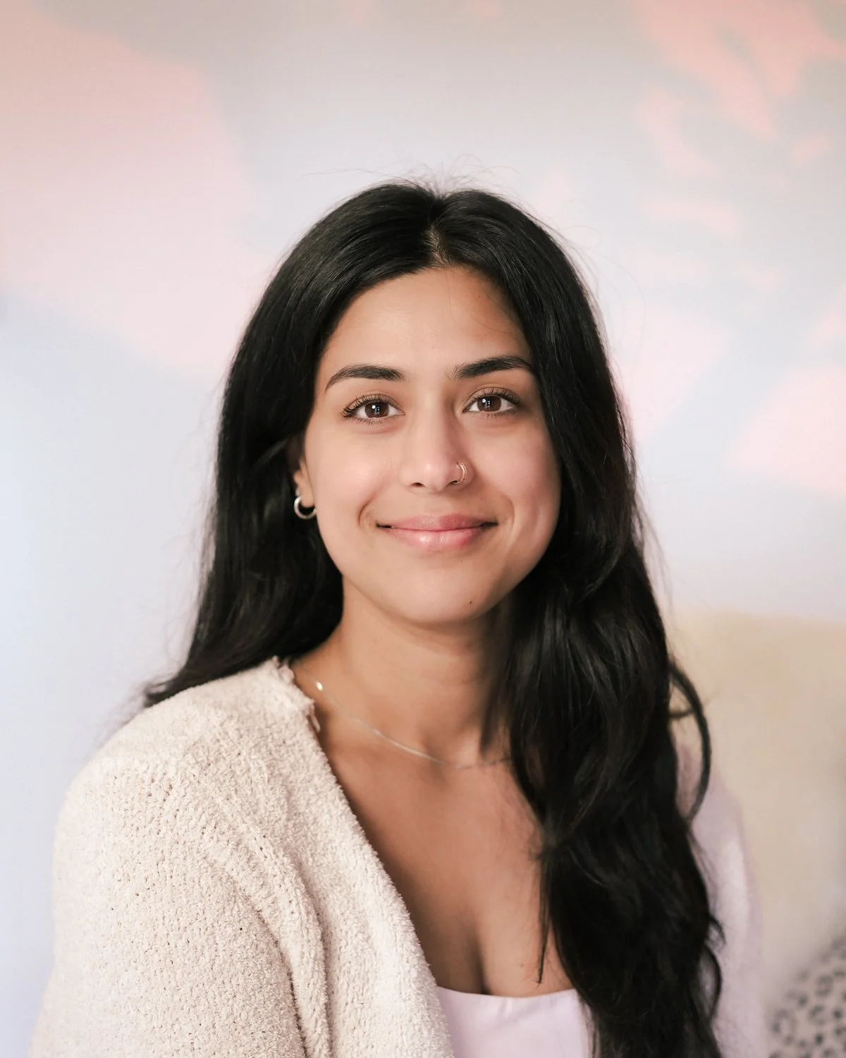 A young woman with long black hair, light skin, wearing a beige sweater, silver earrings, and a nose ring, smiling softly against a soft pastel background.