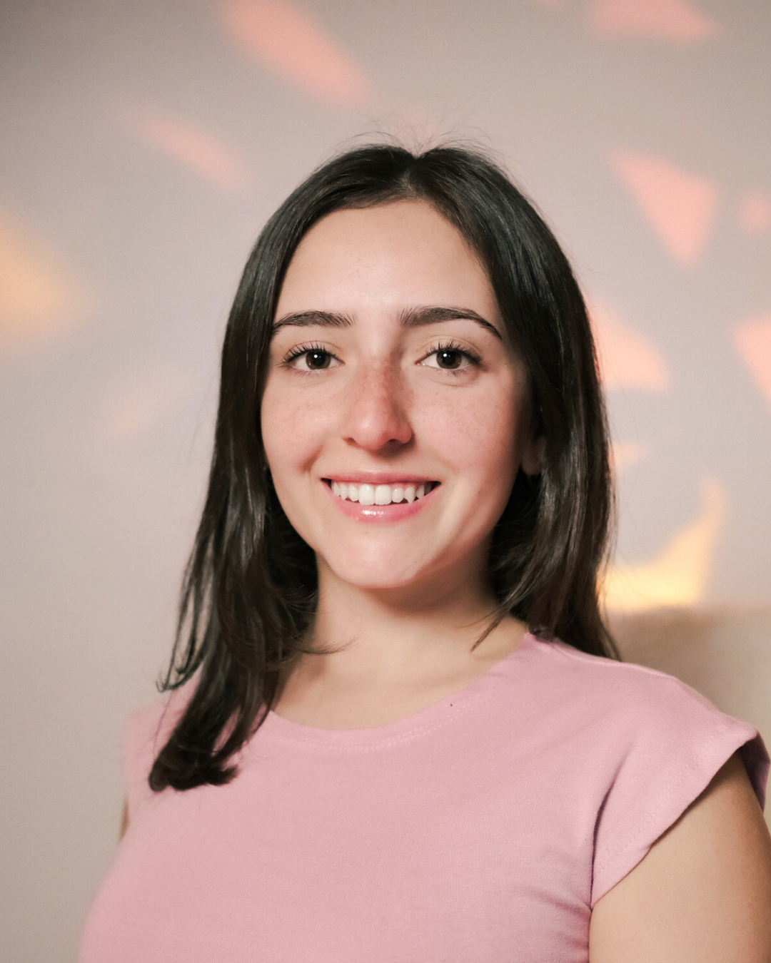 Young woman with brown hair, smiling, wearing a white t-shirt, standing next to a large green Monstera leaf against a white background.