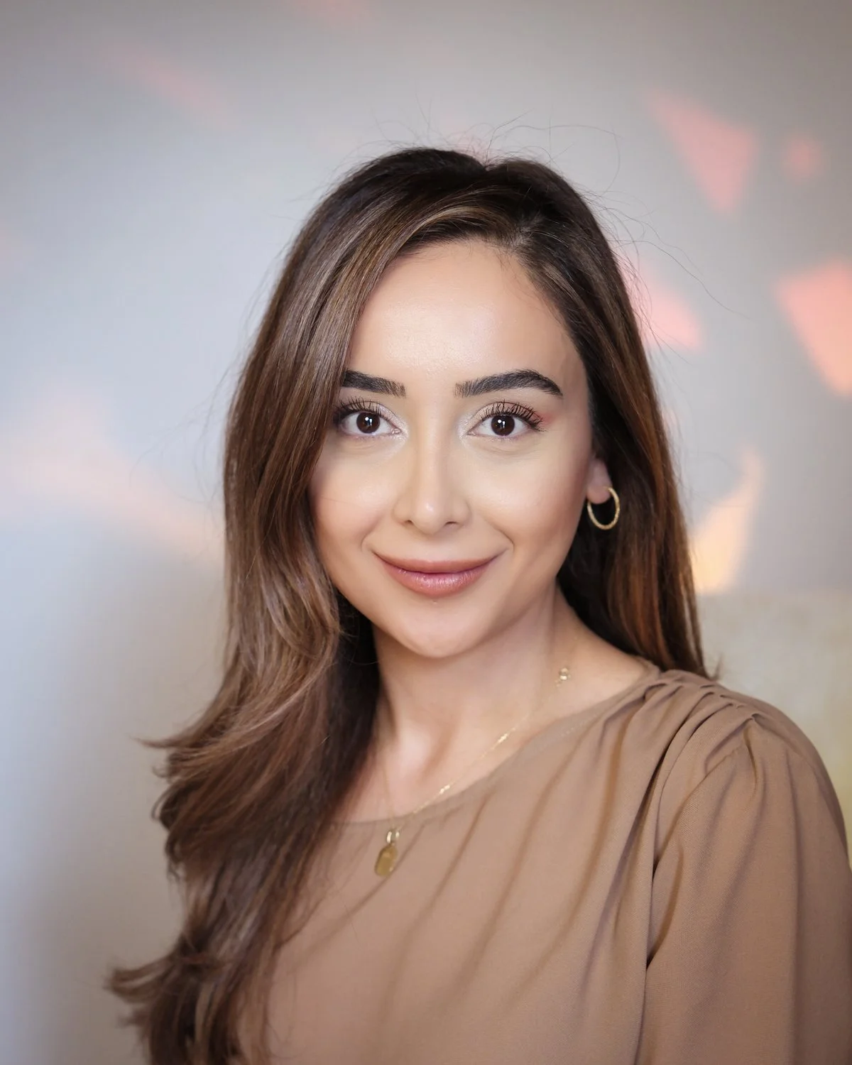 A woman with light skin, long wavy brown hair, and brown eyes, smiling, wearing gold hoop earrings and a beige top, against a blurred colorful background.
