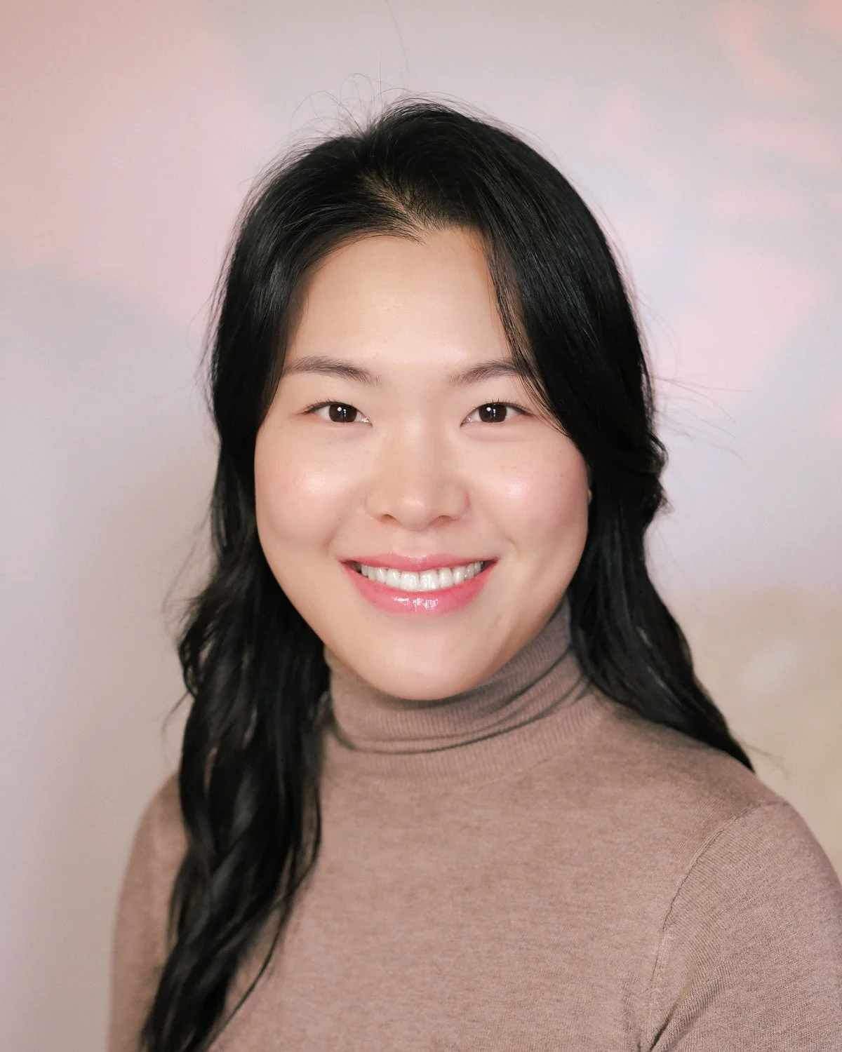 Portrait of an Asian woman with black hair wearing a beige turtleneck sweater, smiling, against a plain light-colored background.