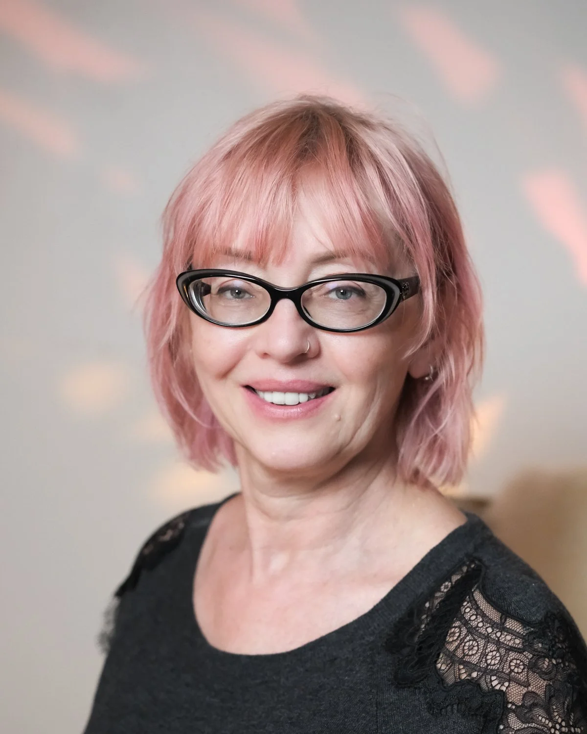 A woman with pink hair, black glasses, and a nose ring smiling at the camera, wearing a black top with lace details on the shoulders.