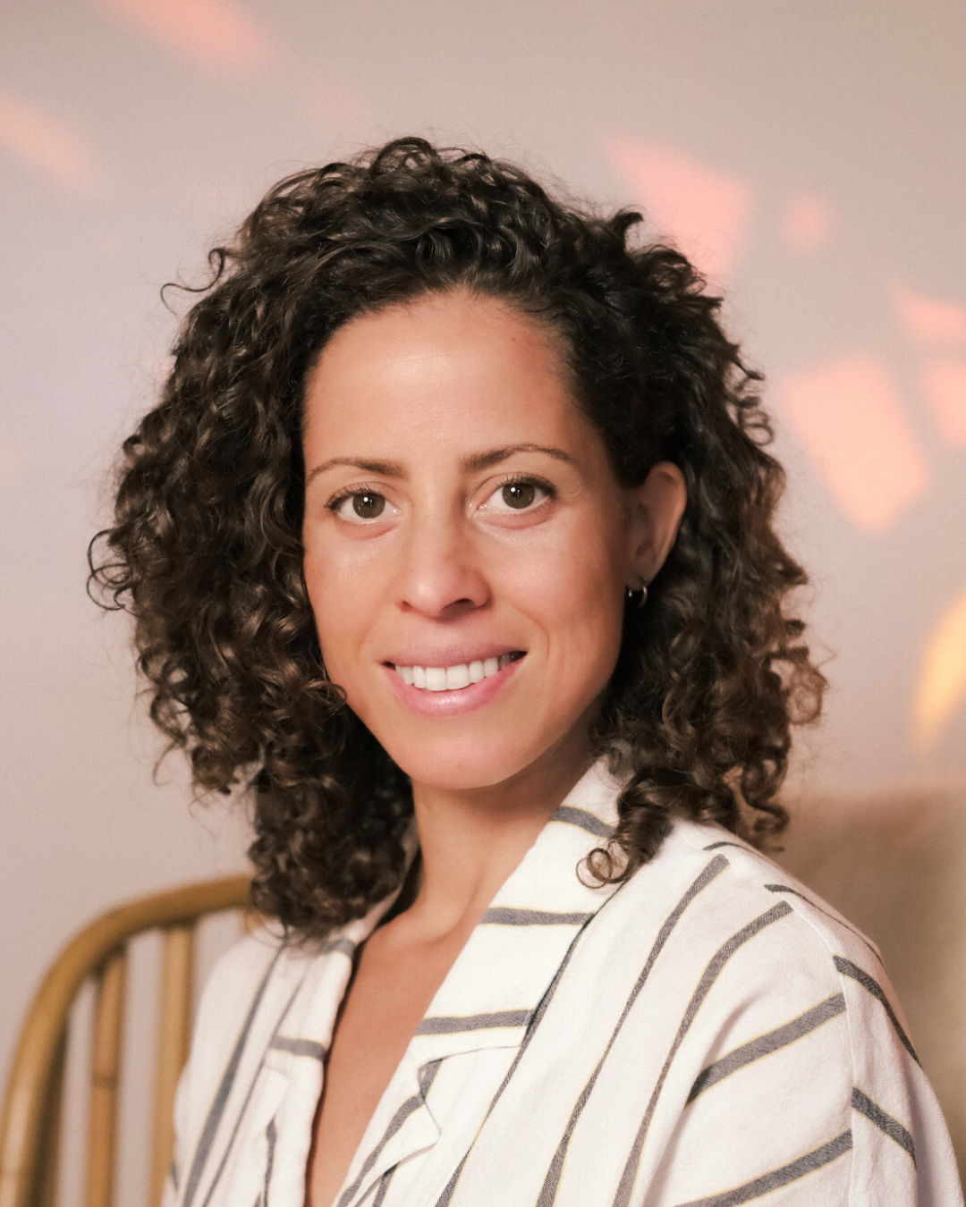 A woman with curly brown hair, wearing a white shirt with black stripes, smiling and looking at the camera. She is in a room with soft pink and orange light reflections on the wall behind her.