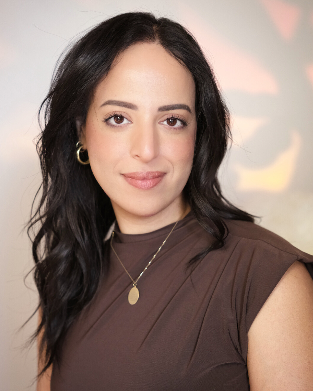 A woman with wavy black hair, brown eyes, and light skin, wearing gold hoop earrings, a gold necklace with an oval pendant, and a brown sleeveless top, standing in front of a softly lit background.