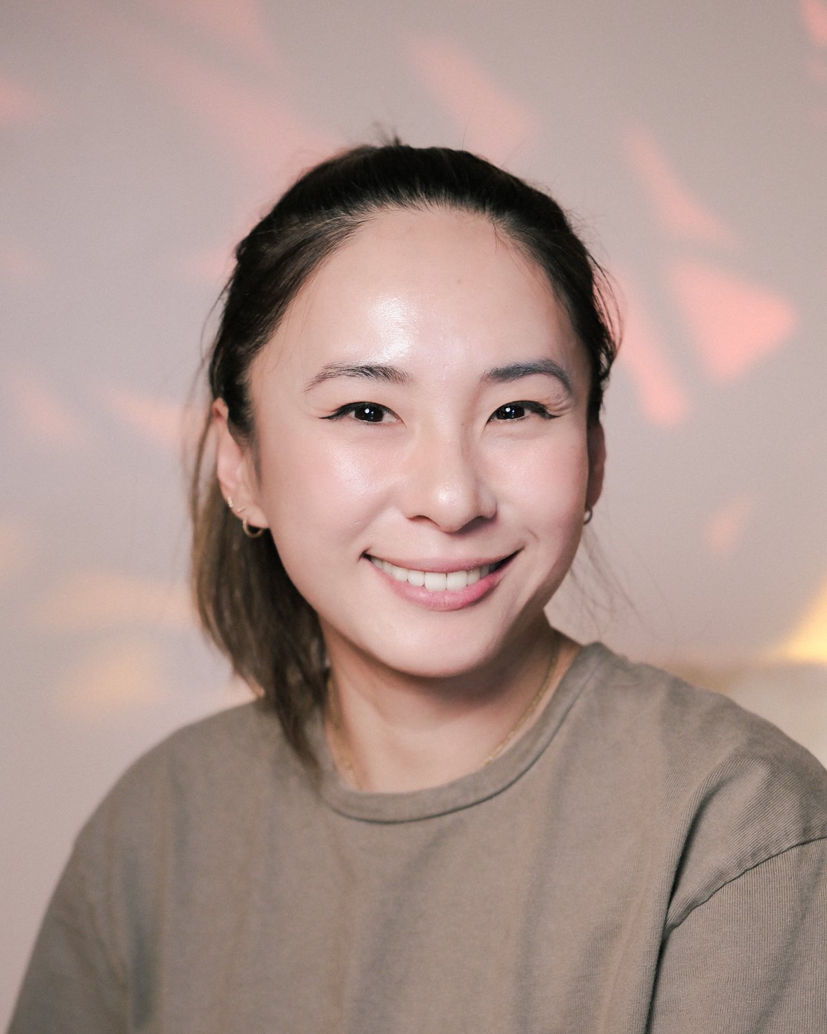 A woman with dark hair tied back, smiling, wearing a beige shirt, with a soft pink and yellow background.
