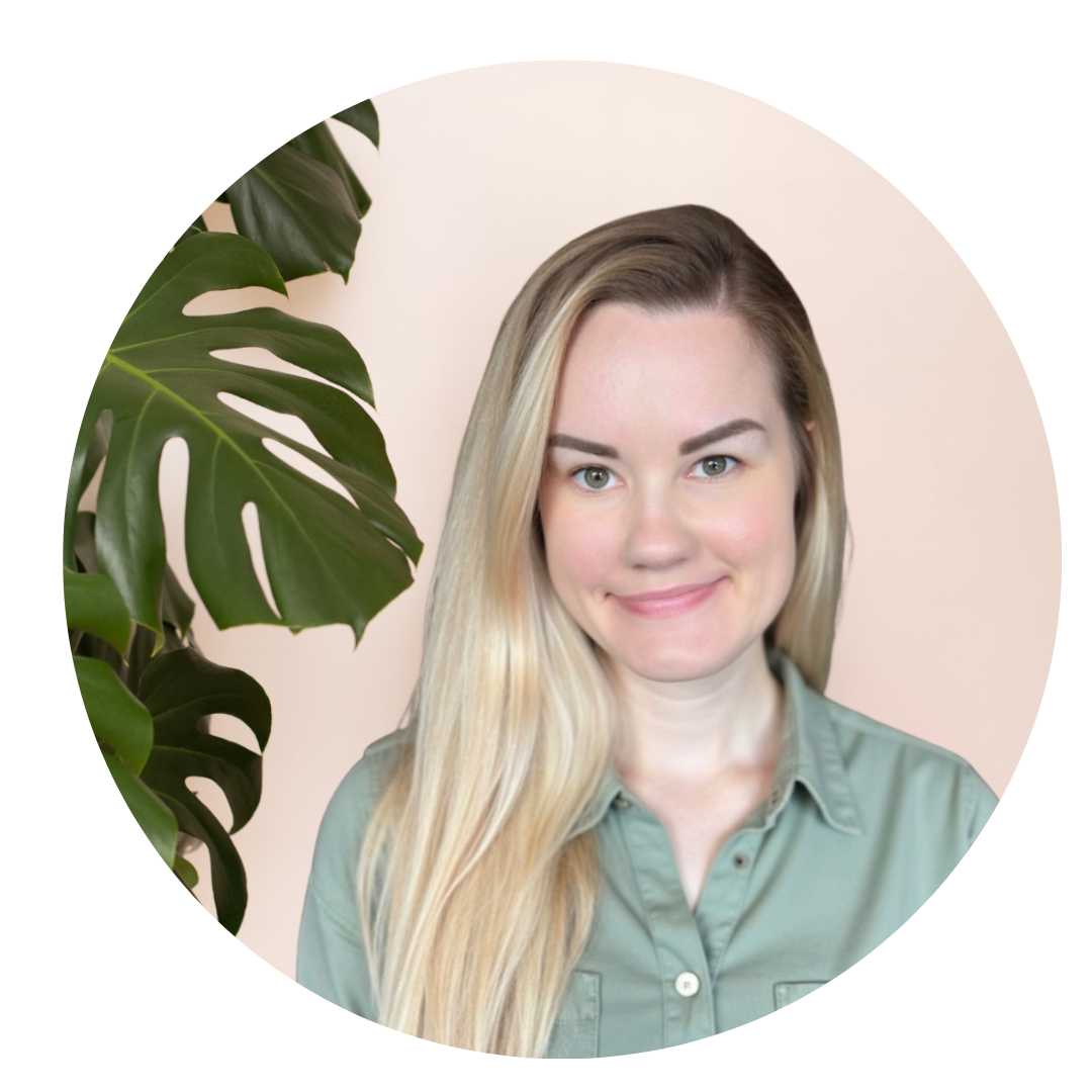 A woman with long light brown hair wearing glasses, a white shirt, and a beige patterned cardigan, standing next to a large green Monstera plant, smiling at the camera.