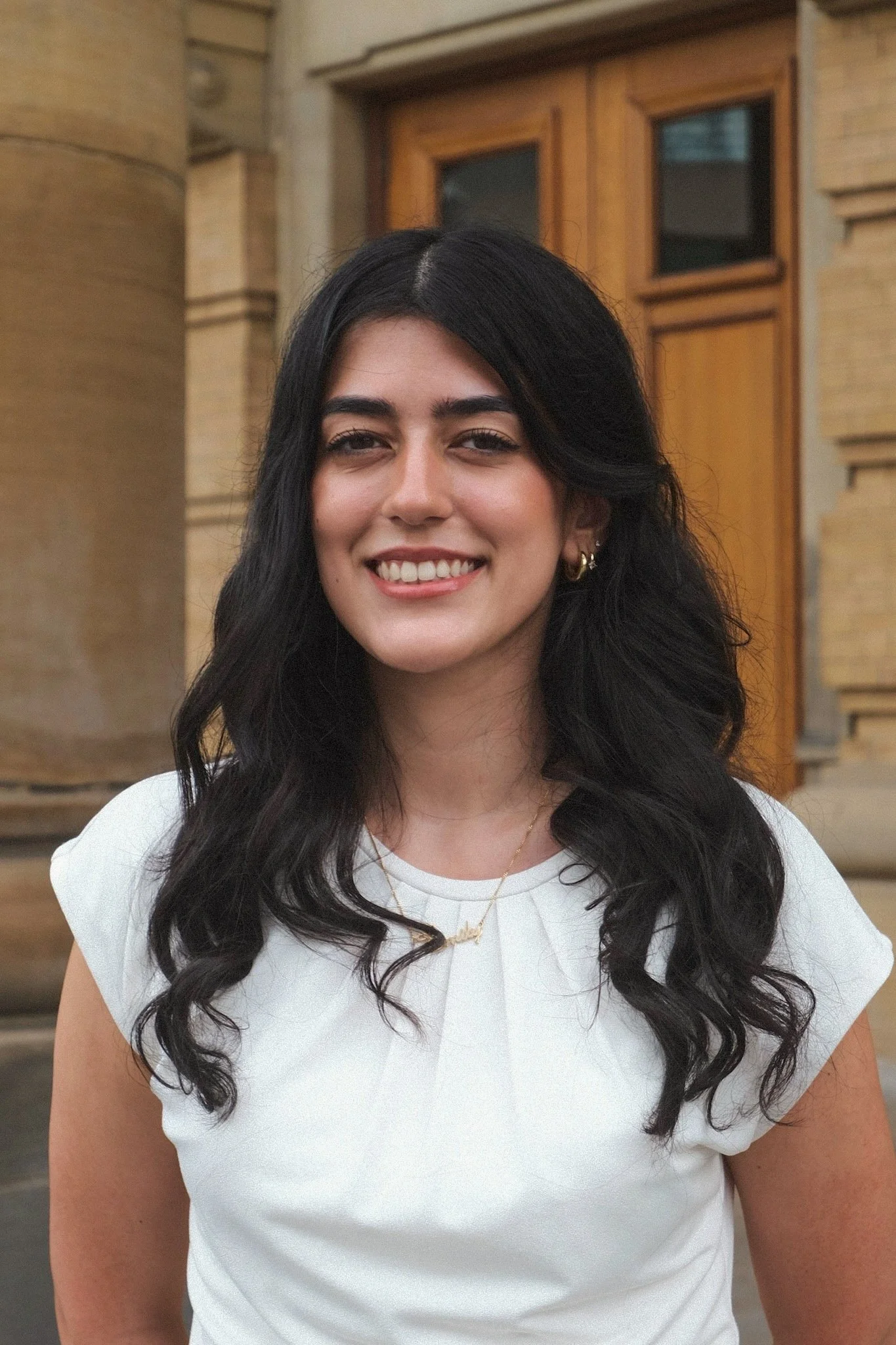A young woman with long dark hair, smiling, wearing a black button-up shirt, standing against a plain white wall.