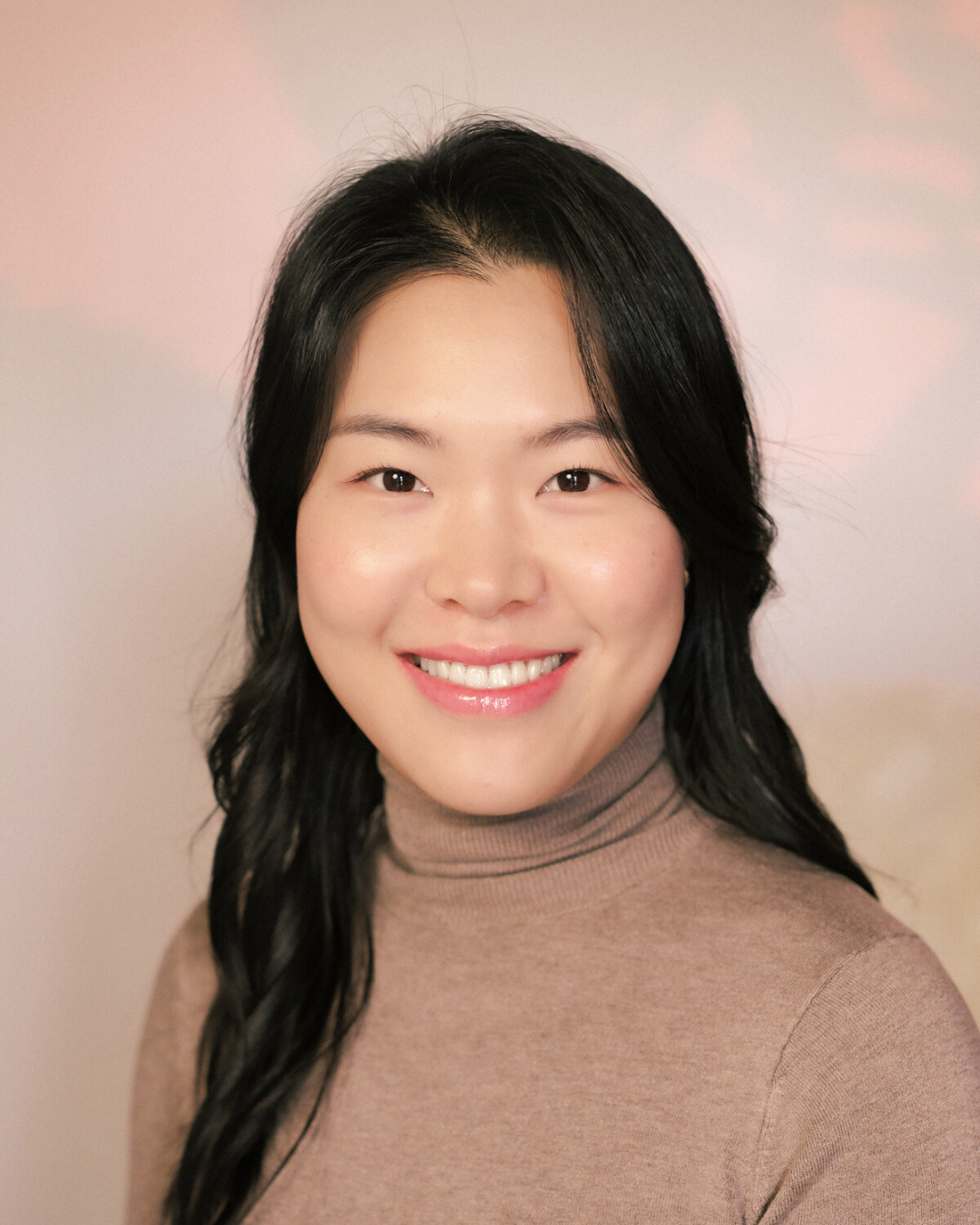 Portrait of an Asian woman with black hair wearing a beige turtleneck sweater, smiling, against a plain light-colored background.