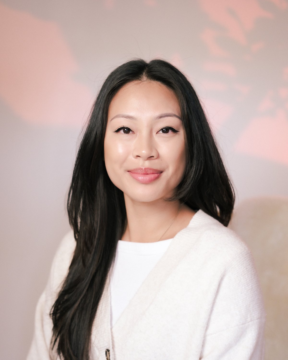 A woman with long black hair, wearing a white top, smiling softly against a pastel pink and beige background.