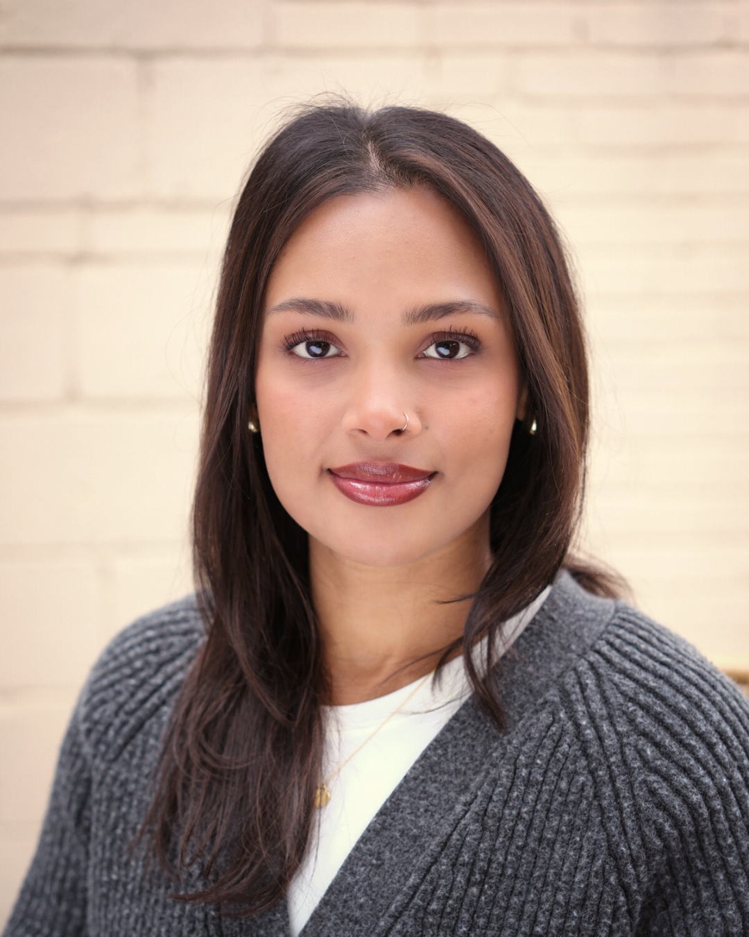 A woman with dark hair and a tan complexion standing with arms crossed, wearing a gray cardigan and white top, in front of a leafy green plant and a plain beige wall.