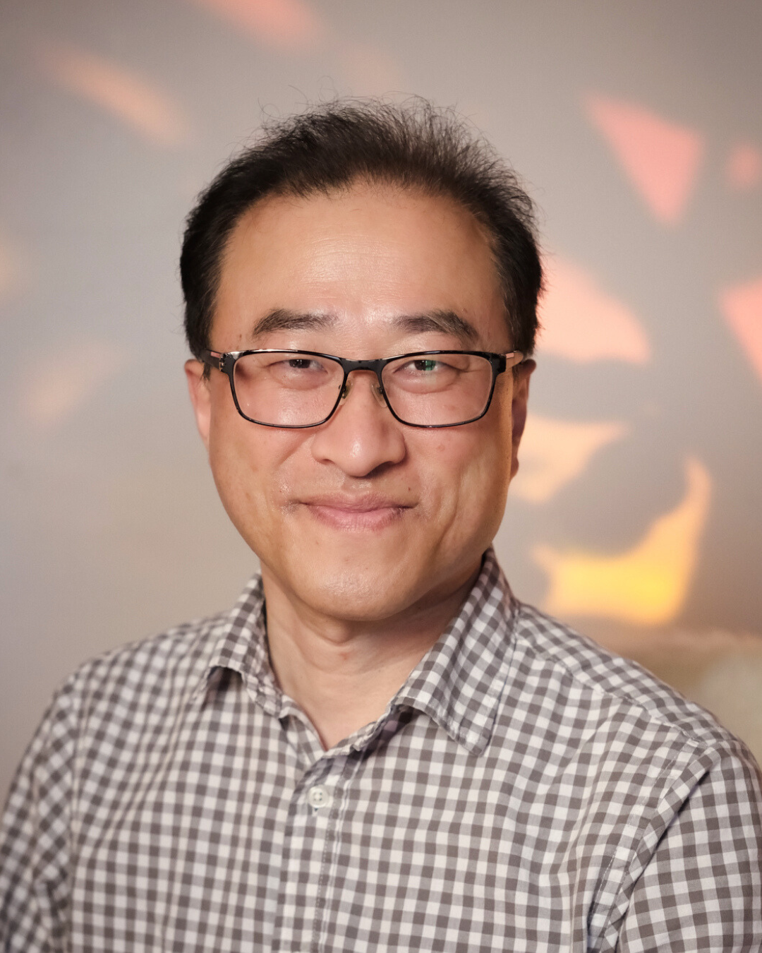 A man with glasses and short black hair, wearing a checkered shirt, smiling at the camera with a blurred background of pink and purple clouds.