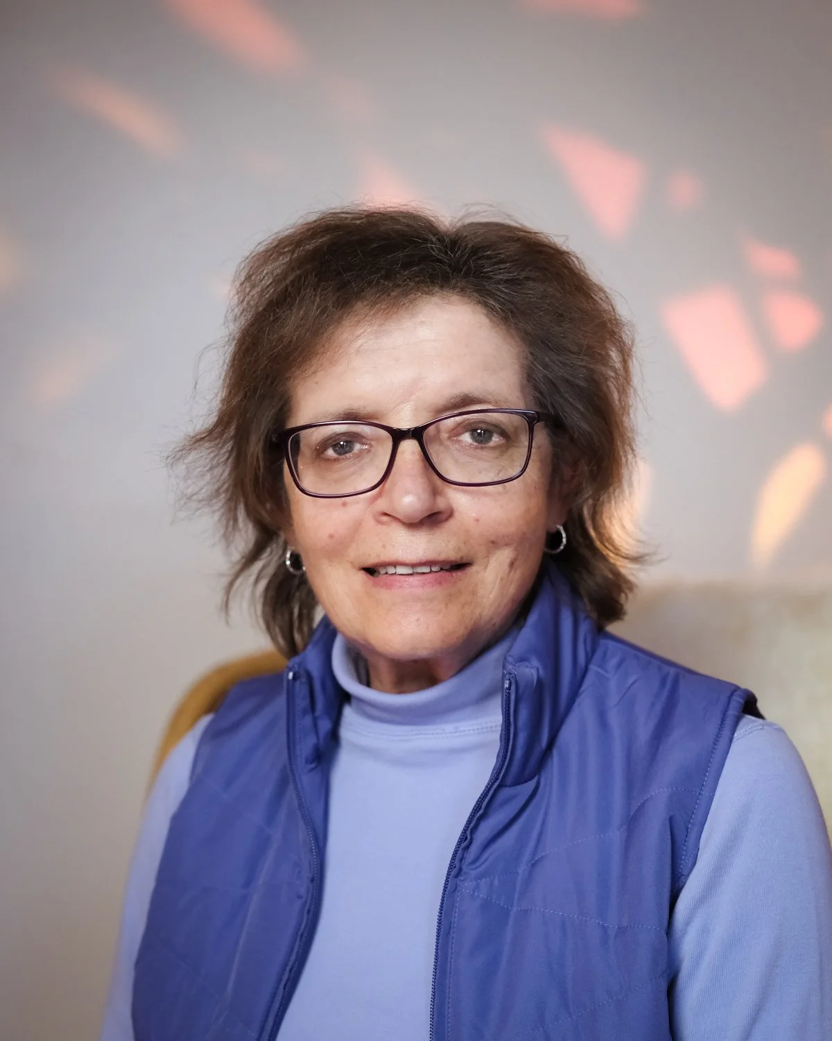 Smiling woman with short brown hair, wearing glasses and a purple turtleneck, standing against a white background.