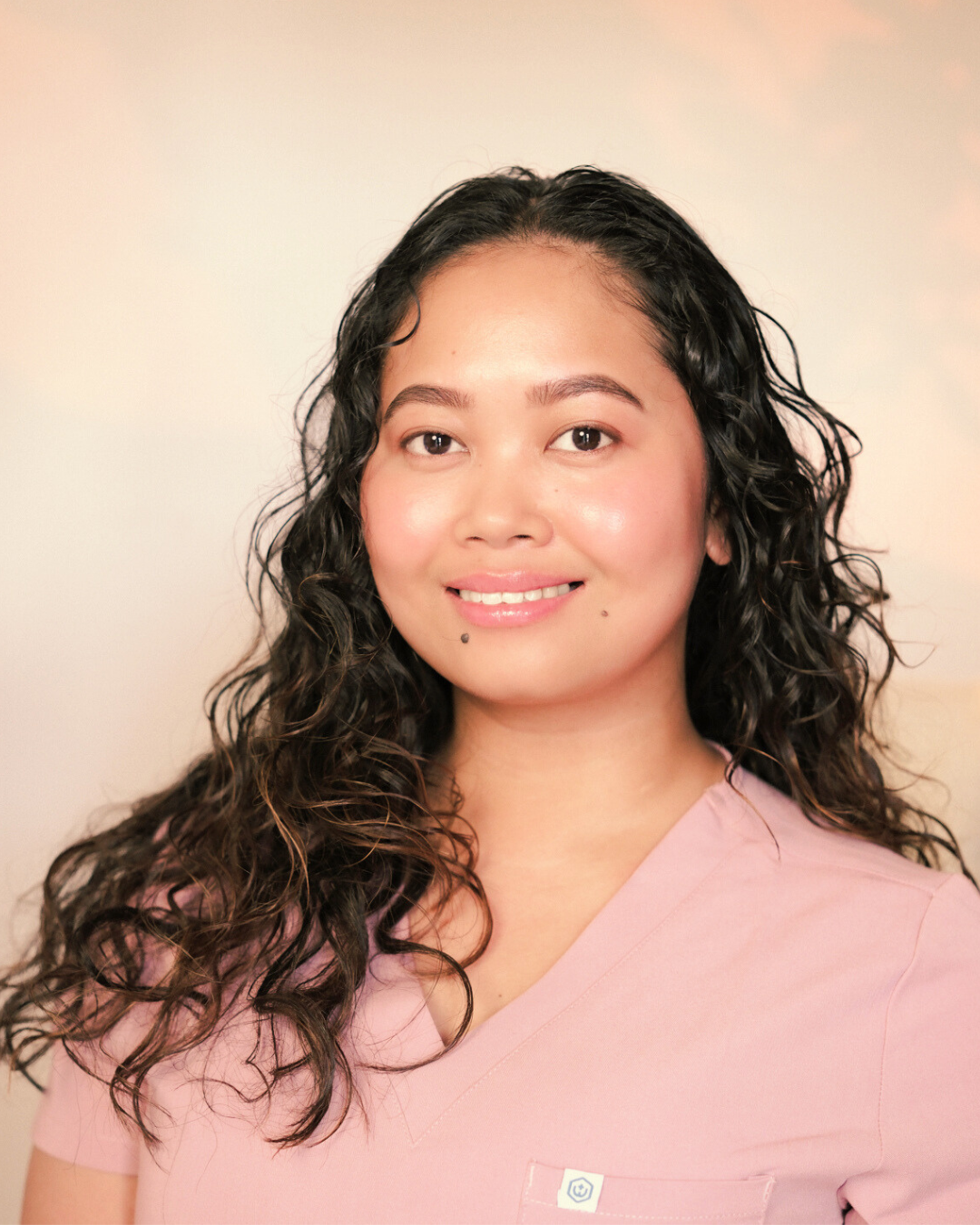A woman with curly black hair and light skin wearing a light pink top, smiling softly against a plain light-colored background.