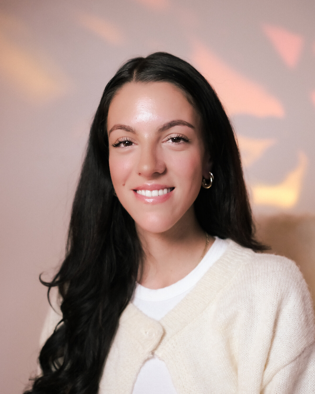 A woman with long black hair smiling at the camera, standing next to a large green monstera leaf, wearing a light blue blouse.