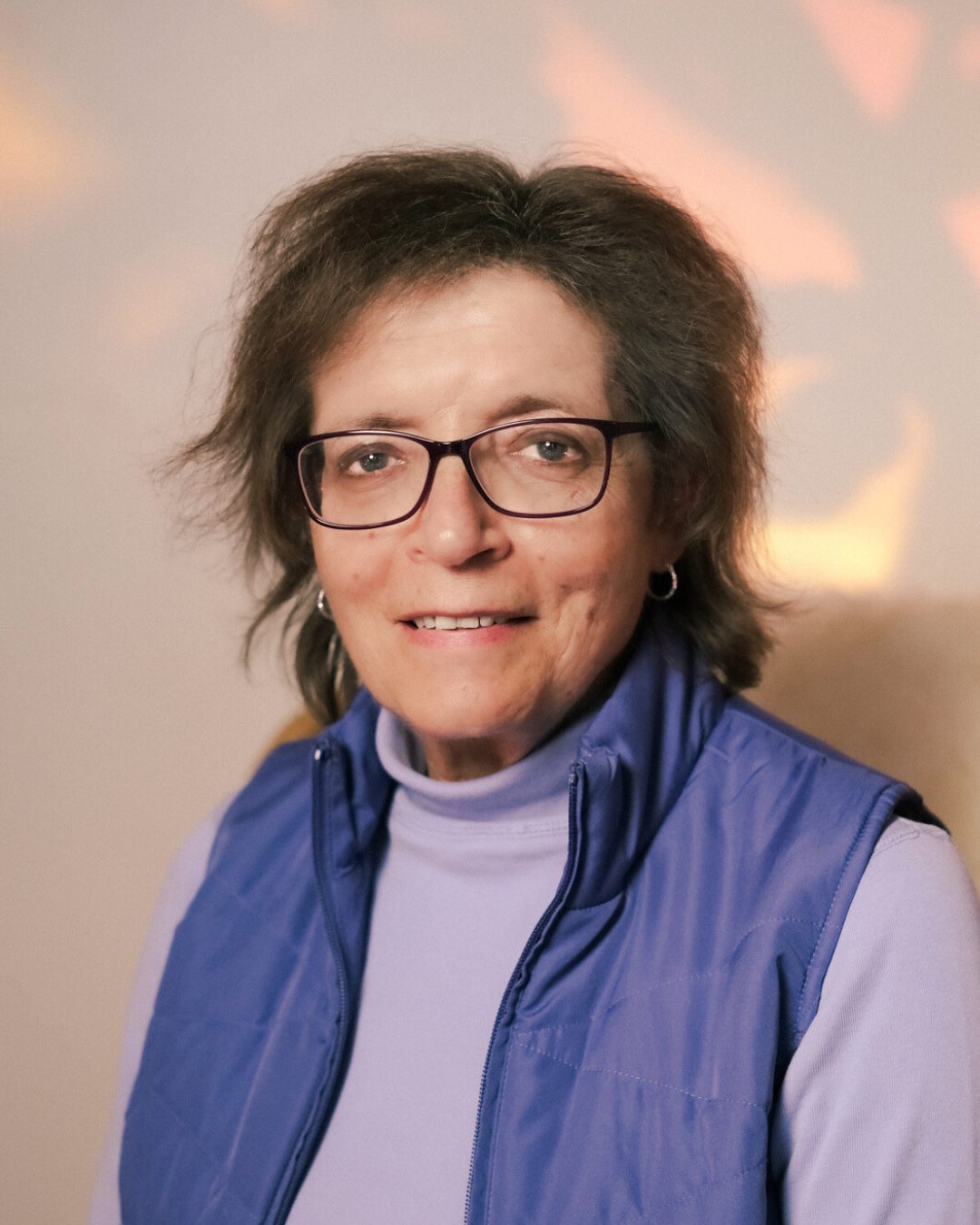 Smiling woman with short brown hair, wearing glasses and a purple turtleneck, standing against a white background.