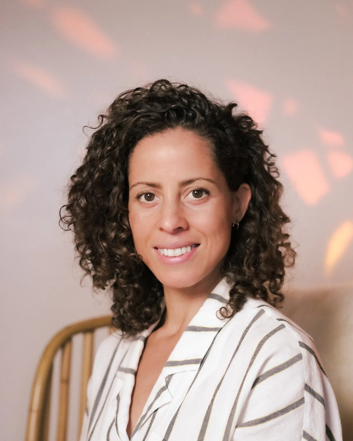 A woman with curly brown hair, wearing a white shirt with black stripes, smiling and looking at the camera. She is in a room with soft pink and orange light reflections on the wall behind her.