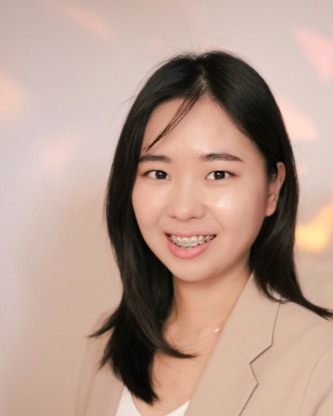 Young woman with long dark hair, glasses, and a gray University of Toronto sweatshirt smiling with arms crossed, standing next to a large green monstera plant against a plain light-colored background.