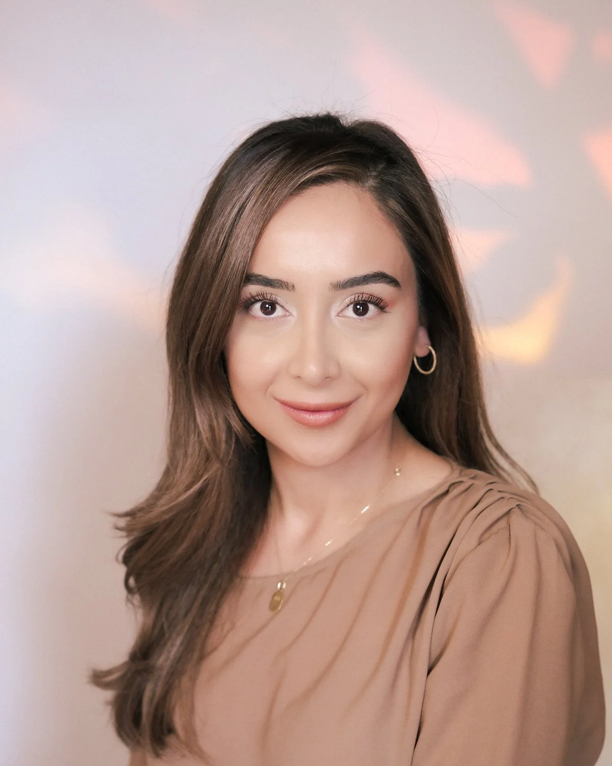 A young woman with long brown hair, wearing a beige top and gold jewelry, smiling at the camera against a soft background with rainbow-like light reflections.