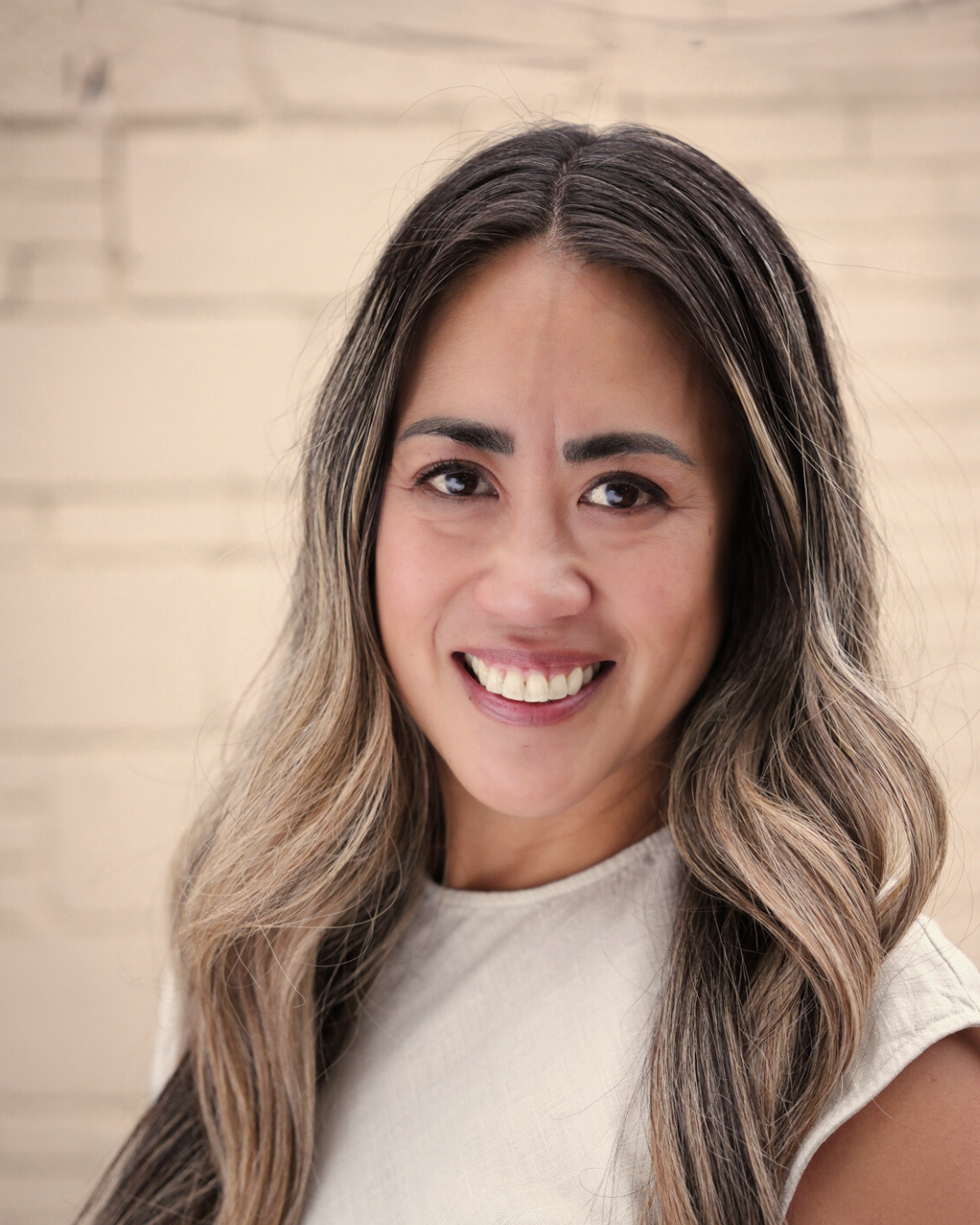 A woman with long wavy hair and a light-colored blazer stands in front of a white background with a large green monstera leaf to her side.