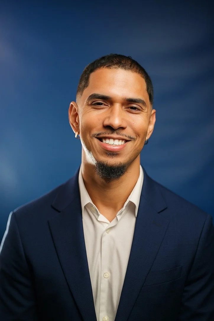 Portrait of a smiling man with short dark hair, a goatee, and a small earring, wearing a navy blue suit and white dress shirt against a dark blue background.