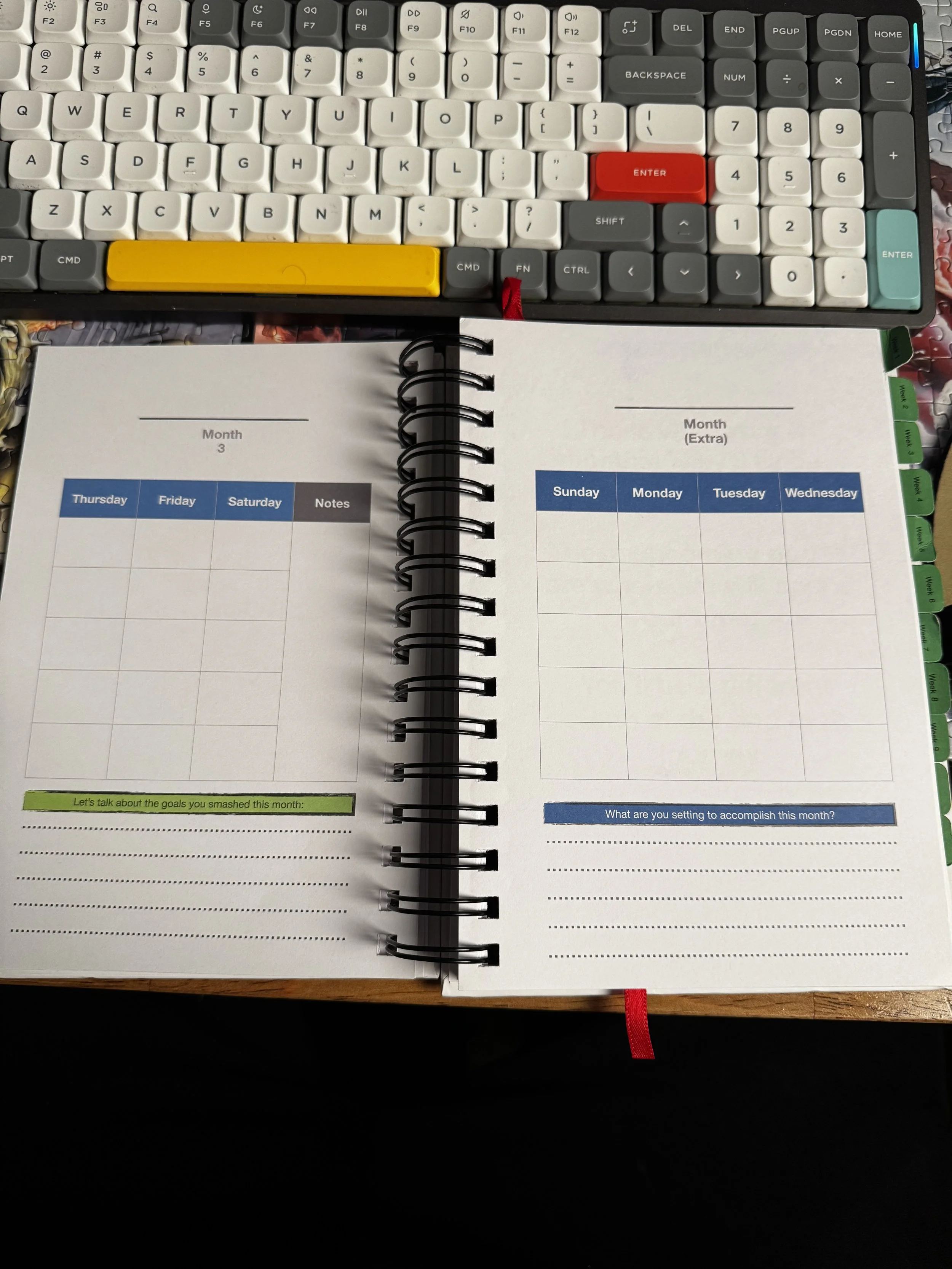 Two-page spread of a desk planner opened to a monthly calendar, with a computer keyboard above. The planner has a weekly layout on the left page for Thursday to Saturday, and an empty weekly layout on the right page for Sunday to Wednesday, with colorful tab dividers on the right edge.