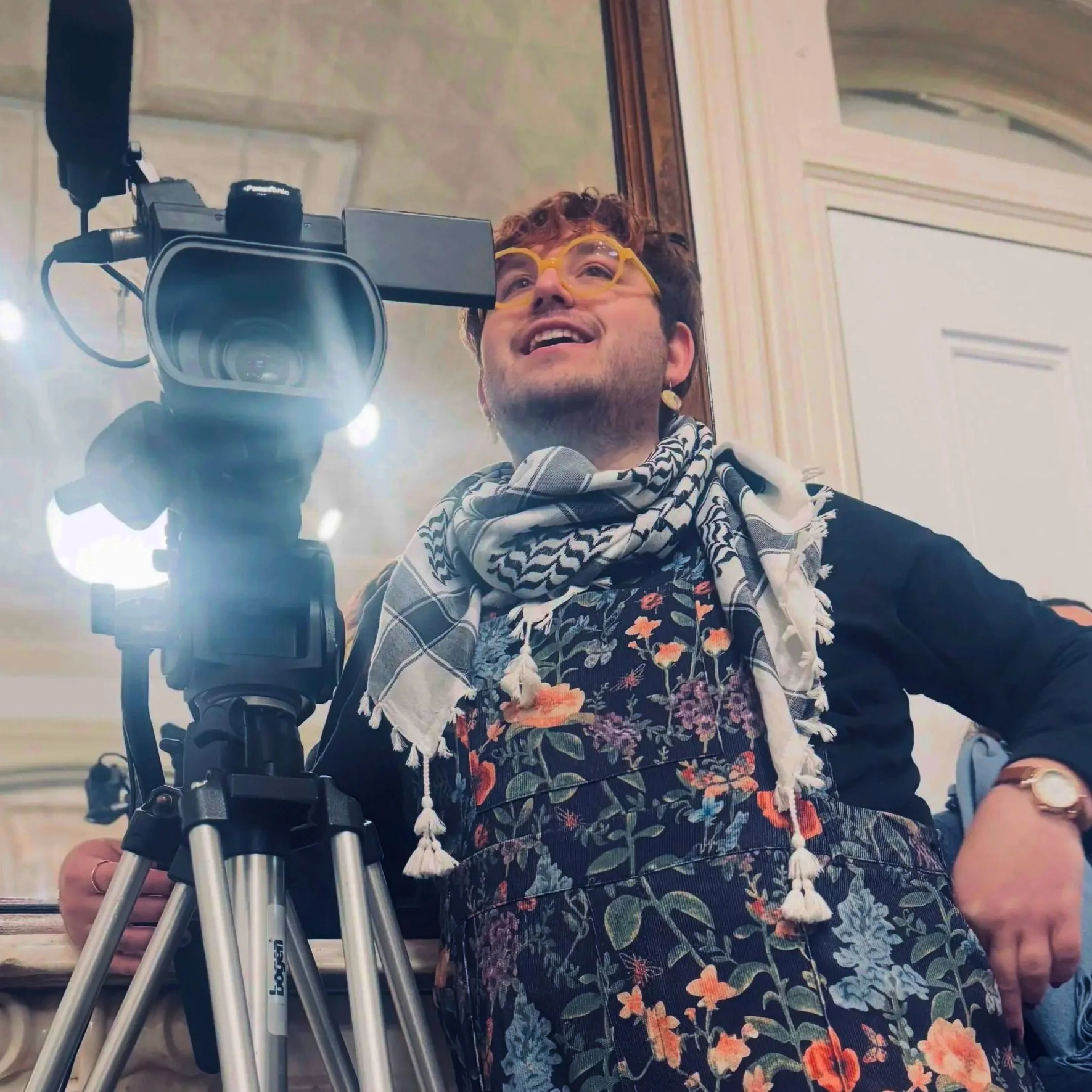Steven with a camera in front of a mirror wearing a floral jumpsuit and a keffiyeh