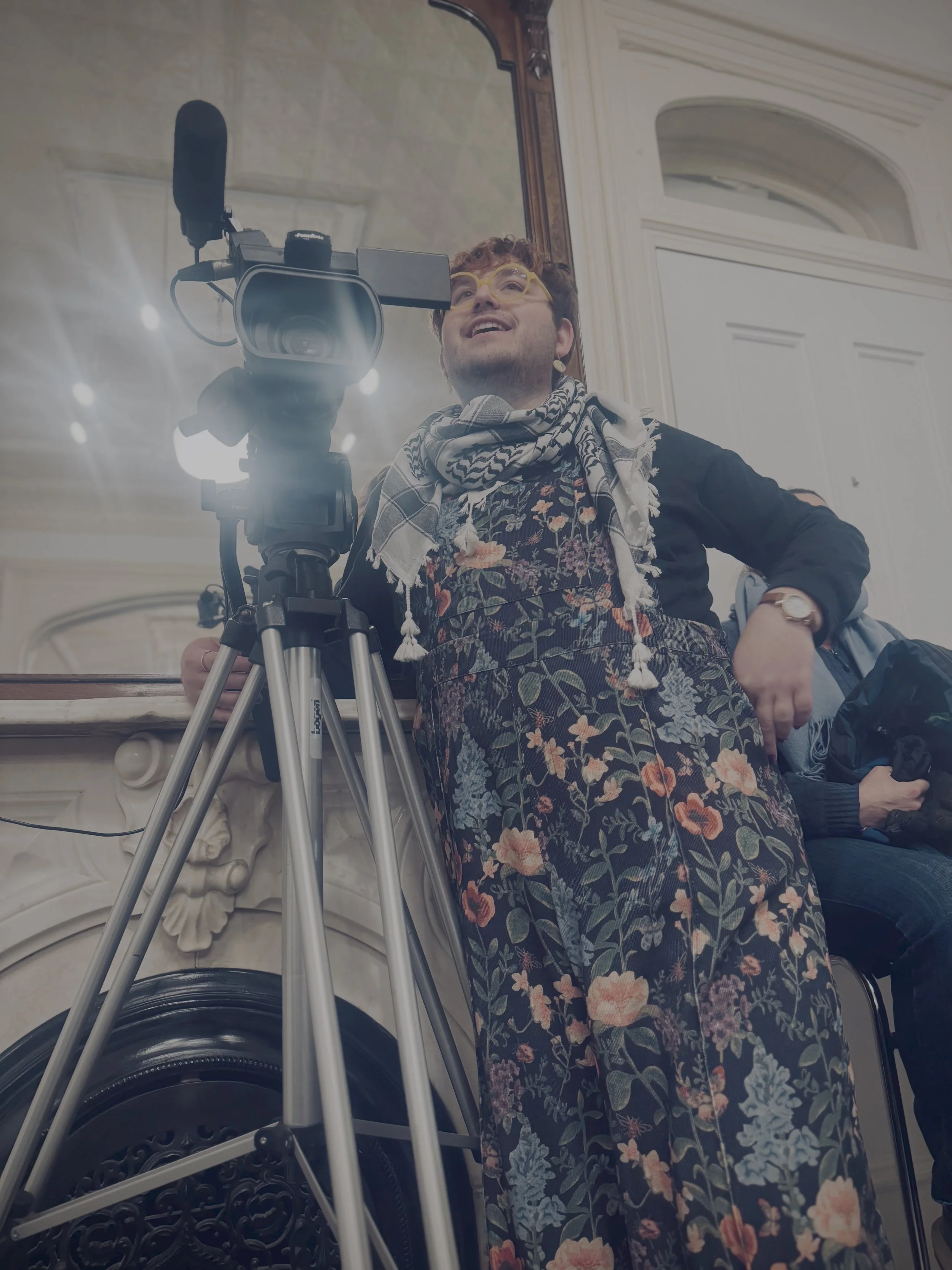 Steven with a camera in front of a mirror wearing a floral jumpsuit and a keffiyeh
